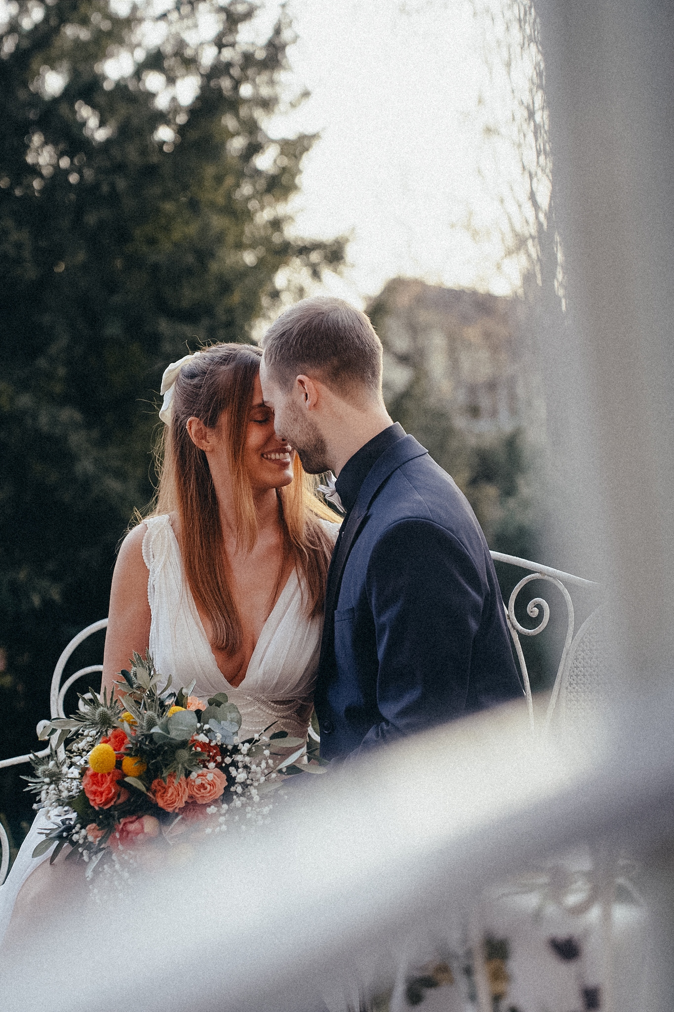 Bride laughs joyfully as groom leans in close on a decorative white bench, holding a bright bouquet.