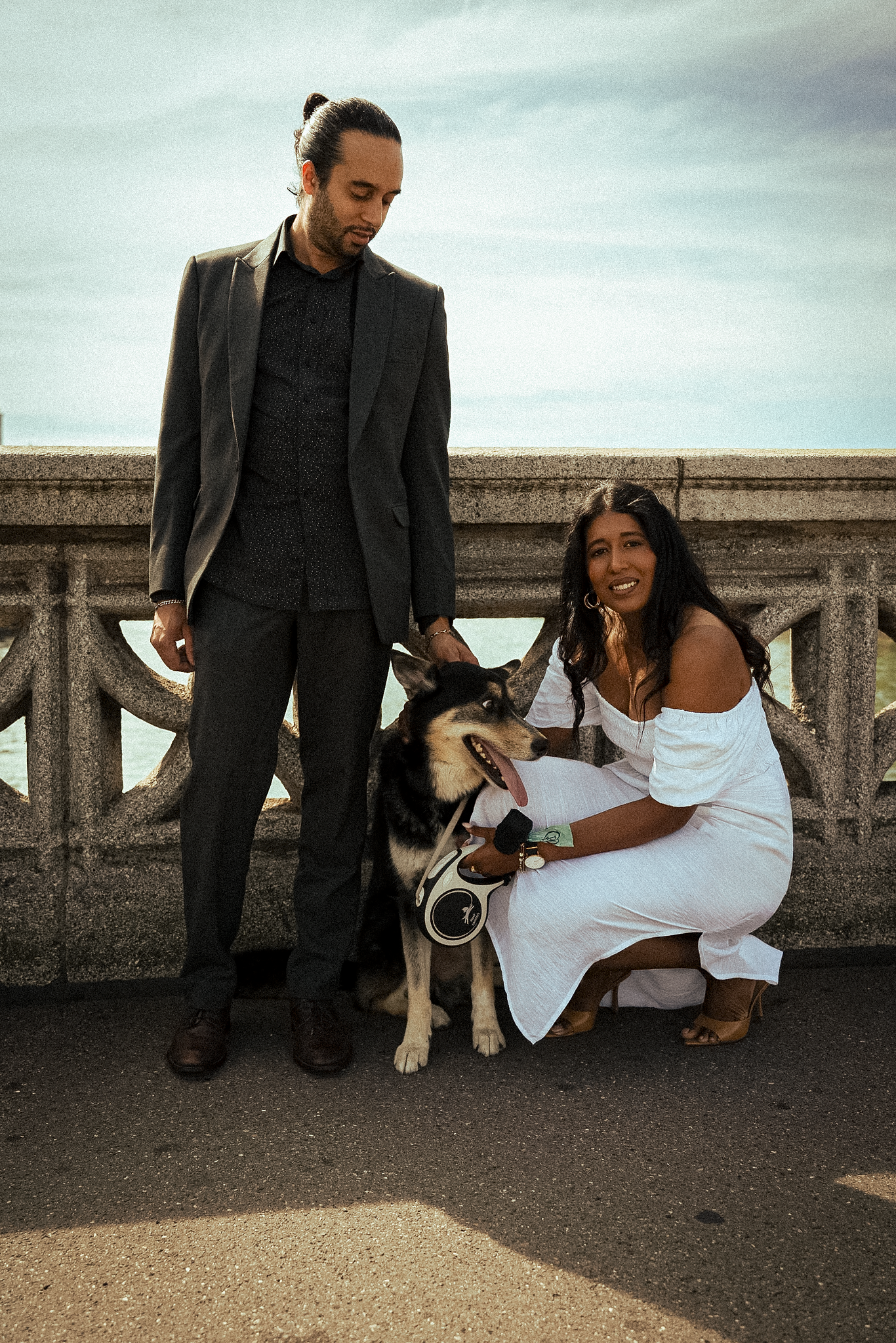 A couple stands on a bridge; the man in a suit and the woman in a white dress smiling at a Husky dog. The tone is relaxed and cheerful.