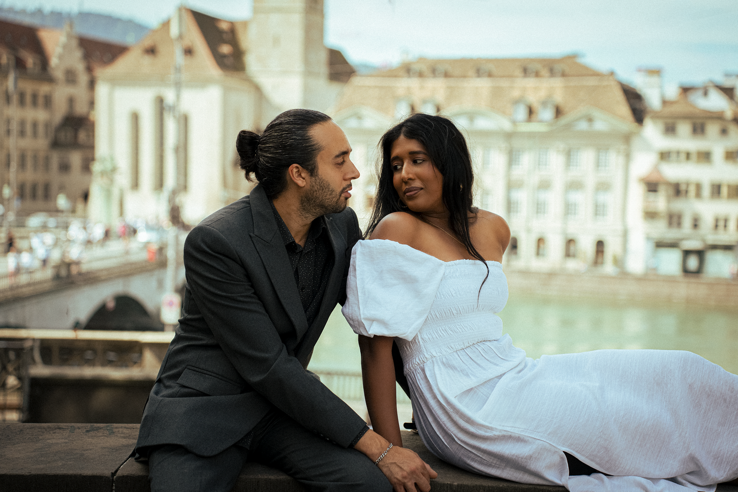 Couple in elegant attire sitting together by Zurich waterfront with historic buildings in background.