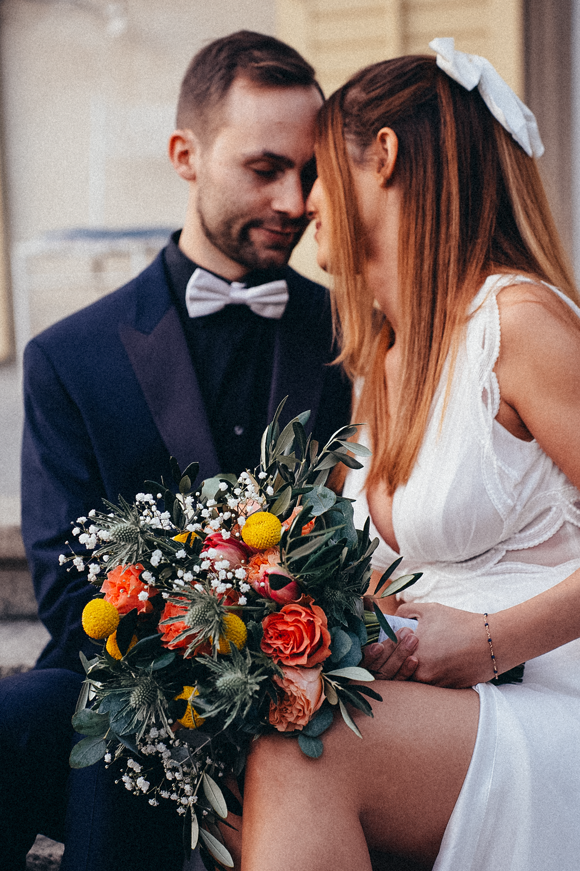 Bride and groom leaning close together, holding a lush bouquet of orange roses and yellow flowers.