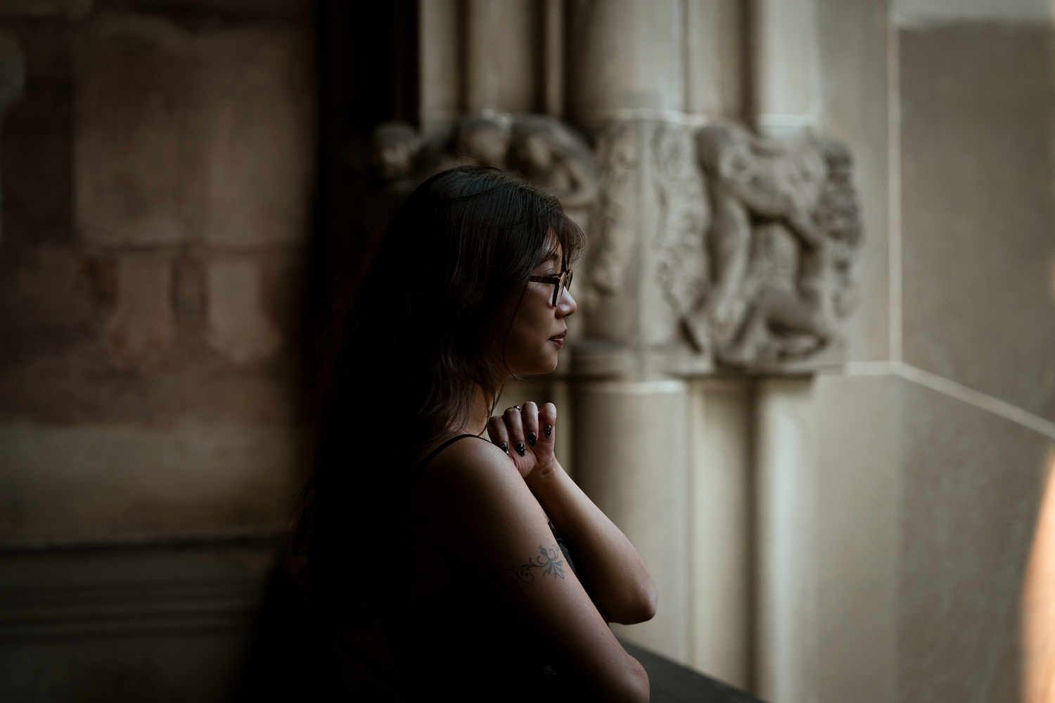 A young woman with glasses prays with hands clasped near ornate stone carvings in a dimly lit historic building.