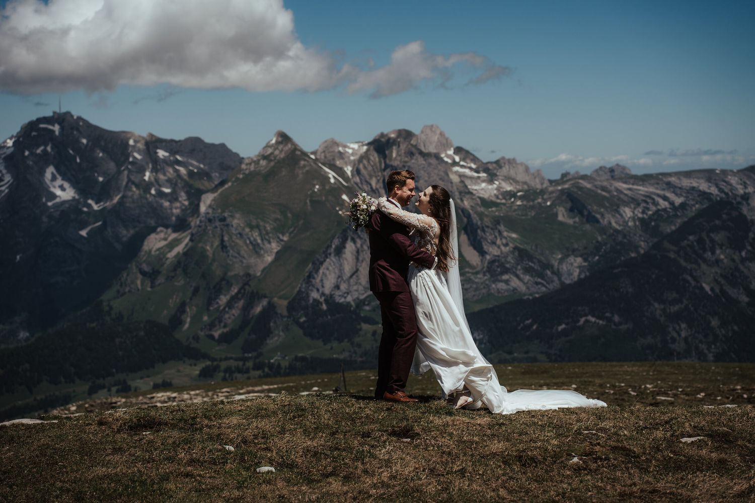 Couple embracing in wedding attire on a mountain summit with dramatic Alpine peaks and blue sky backdrop.