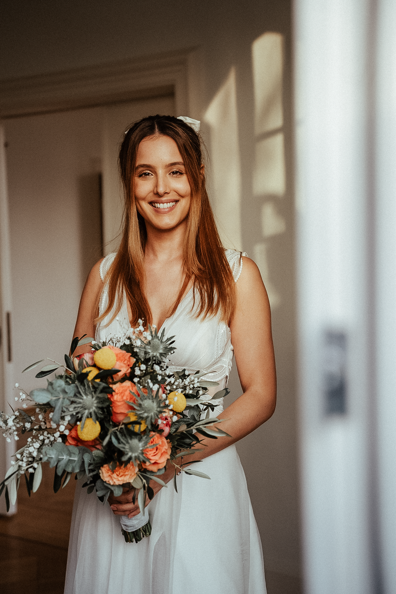 Radiant bride in white dress smiles warmly indoors, holding a beautiful orange and yellow floral bouquet.