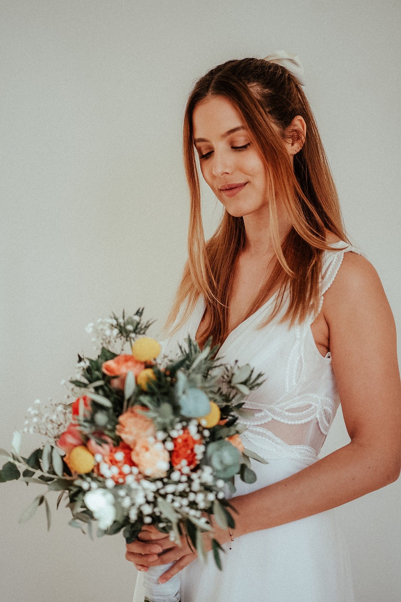 Bride in white dress holding colorful wedding bouquet with roses and greenery, smiling softly against light background.