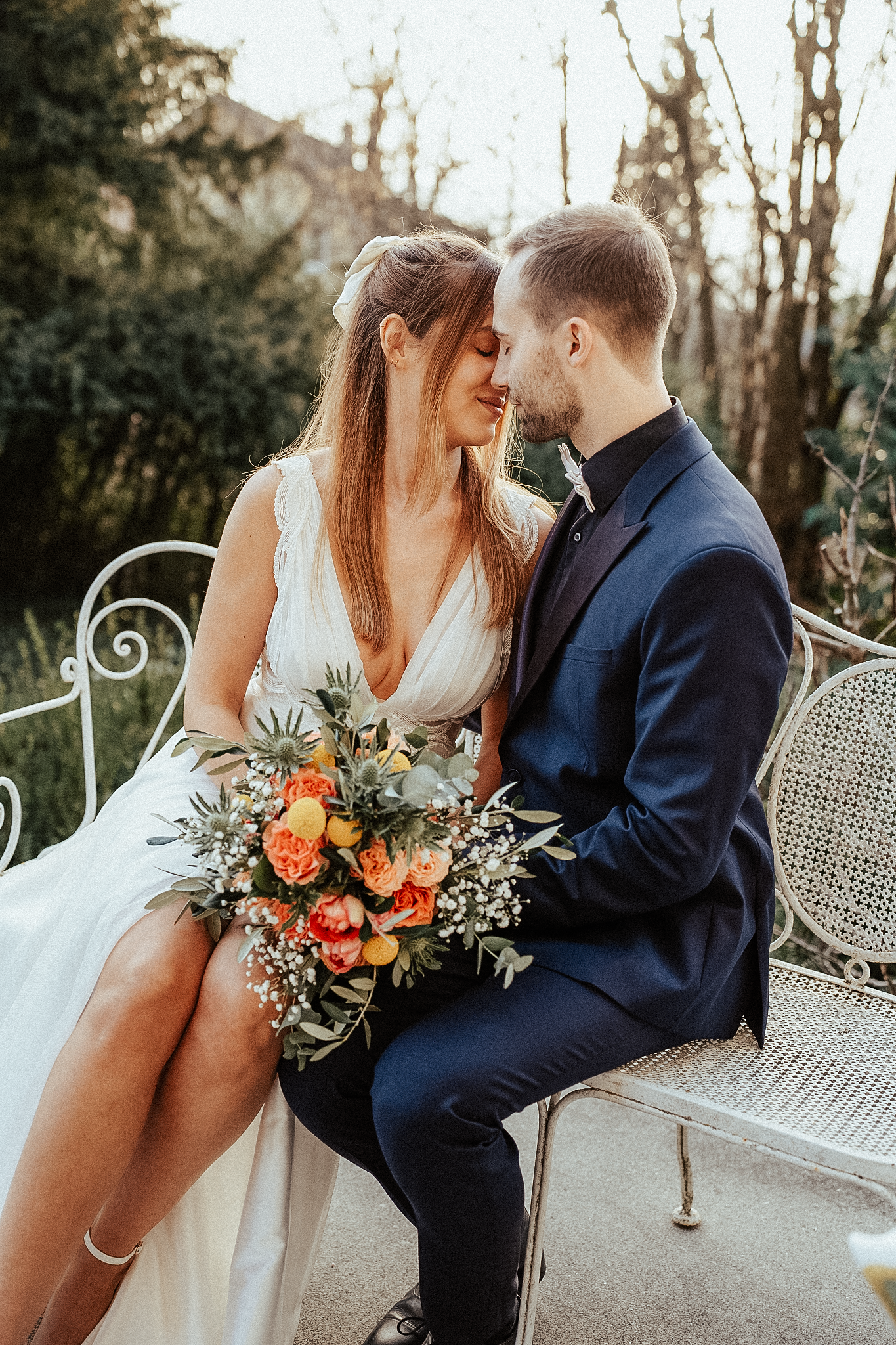 Romantic newlyweds lean in for a kiss on white iron garden bench holding colorful orange bridal bouquet.
