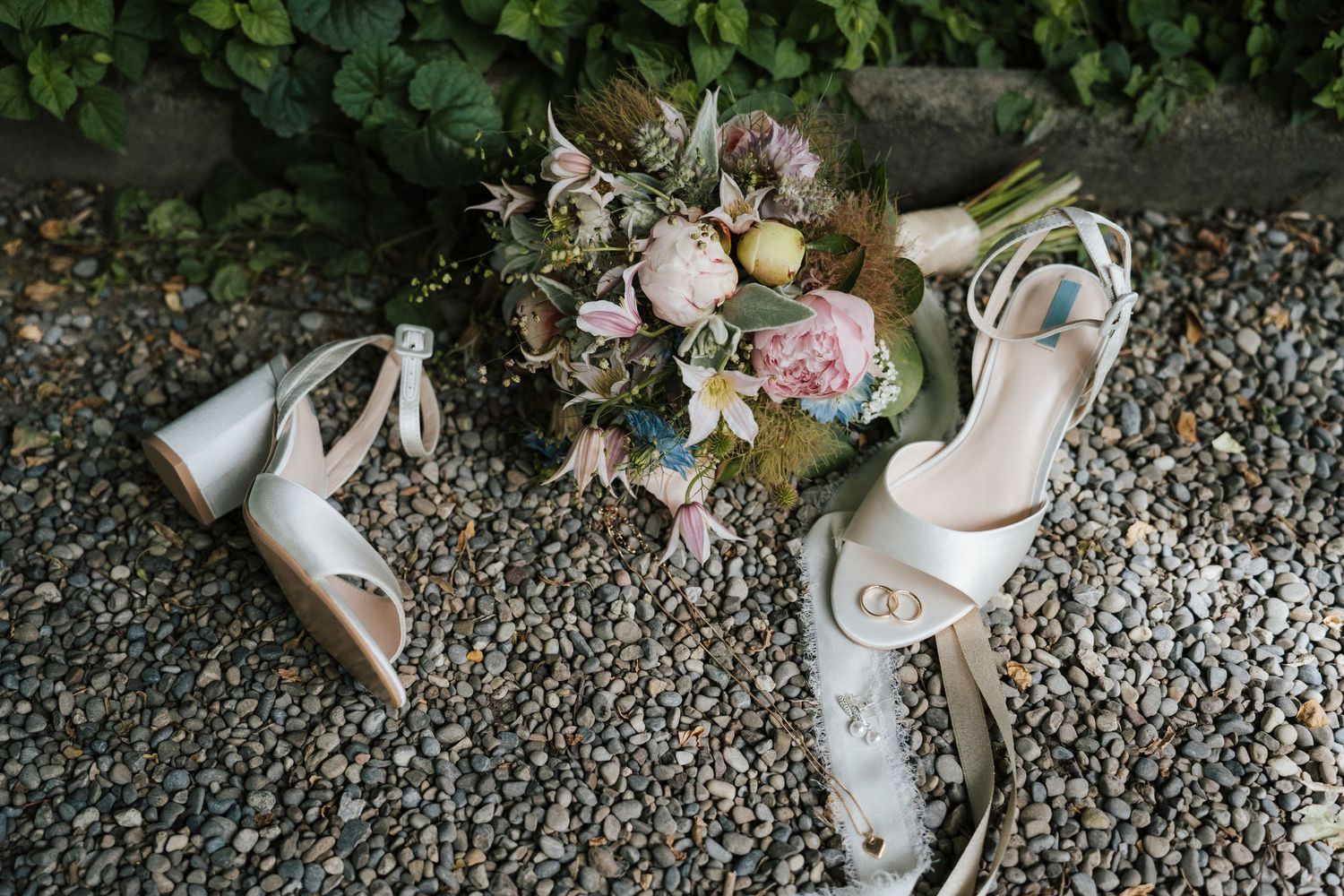 White bridal heels with ribbon ties beside a pastel wedding bouquet of pink peonies on a stone gravel surface.