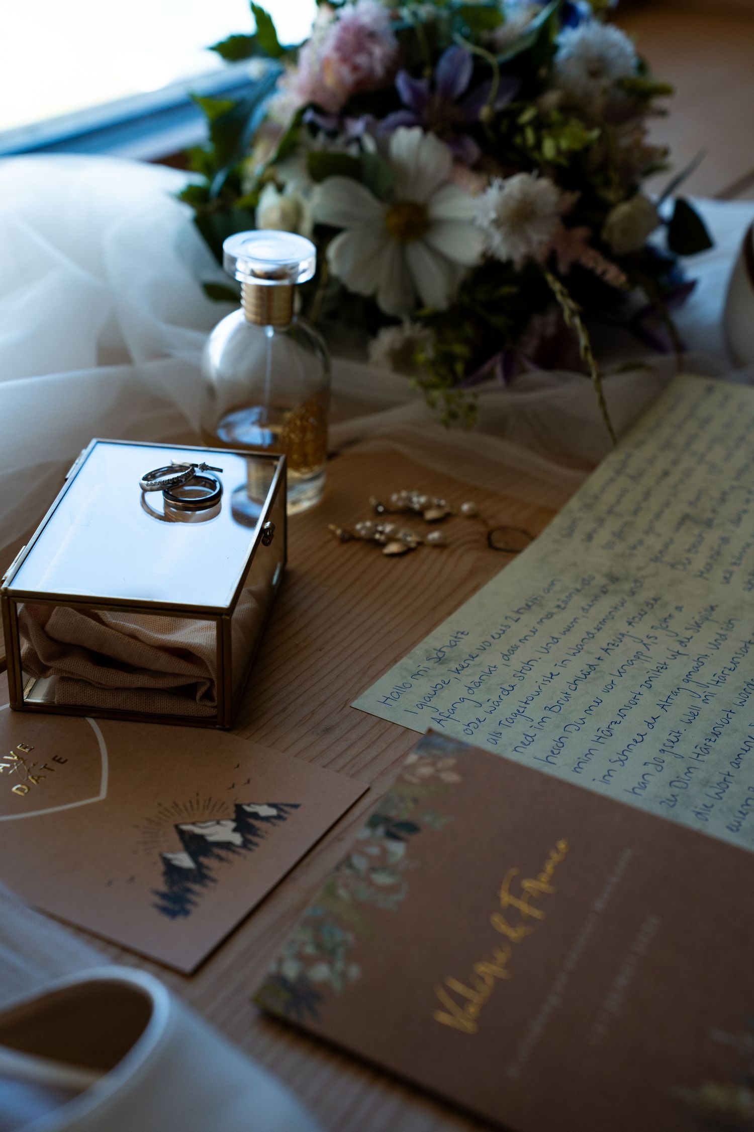 Wedding flat lay with rings, invitation cards, jewelry, perfume bottle, and floral bouquet on a wooden surface.