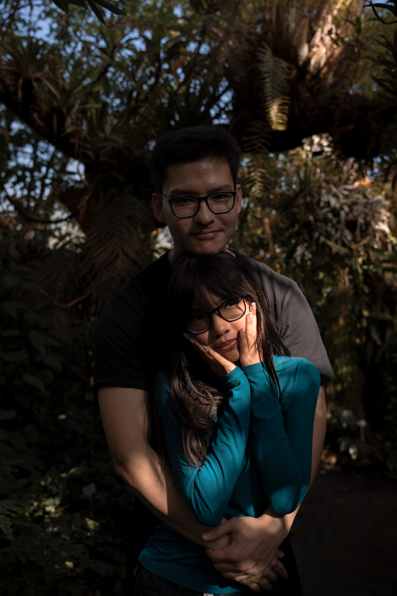 A couple embraces tenderly in a lush garden, the man hugging the woman from behind as she rests her face on her hands.