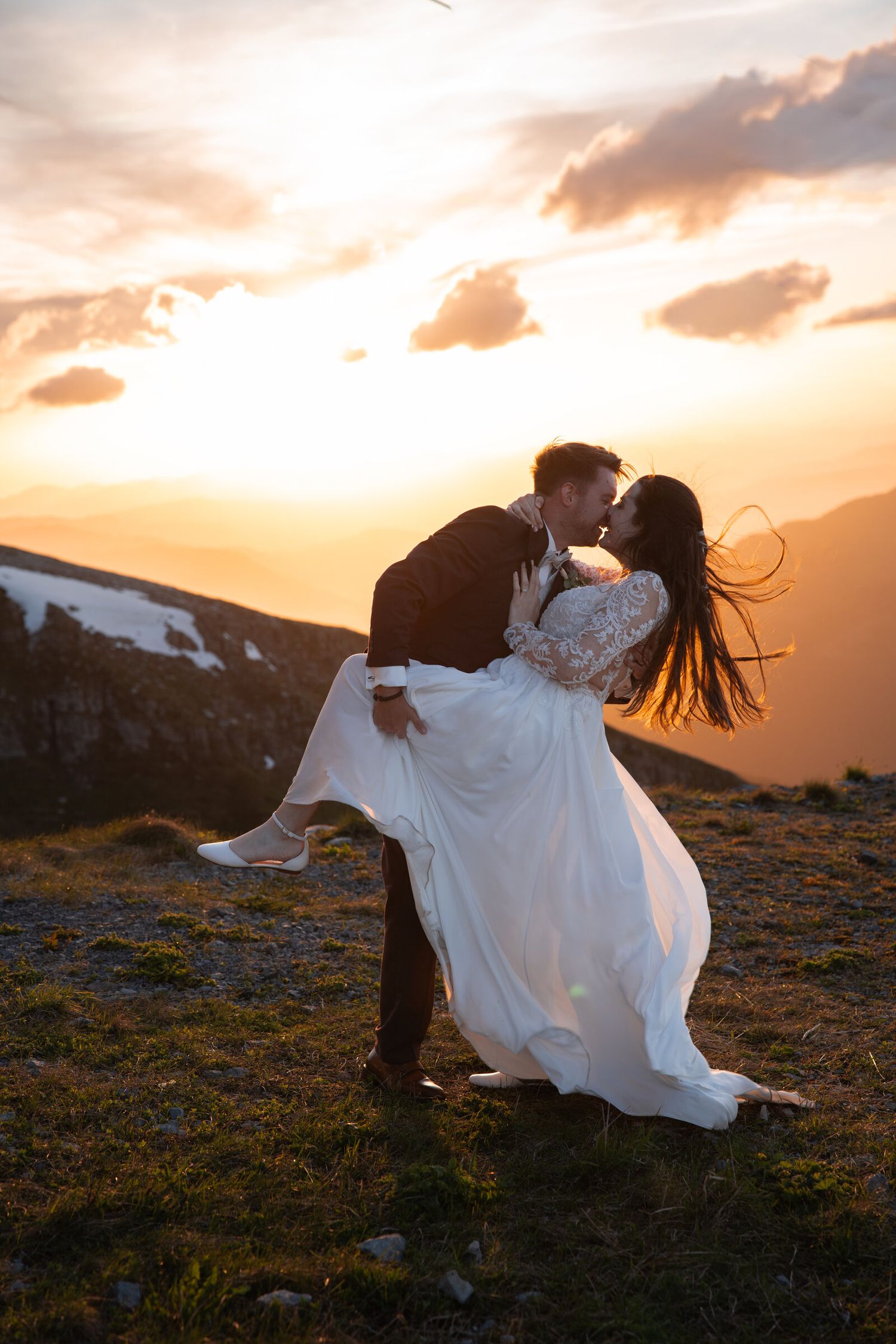 Groom lifting bride in white dress while kissing at sunset on a mountain, with golden sky and clouds behind them.