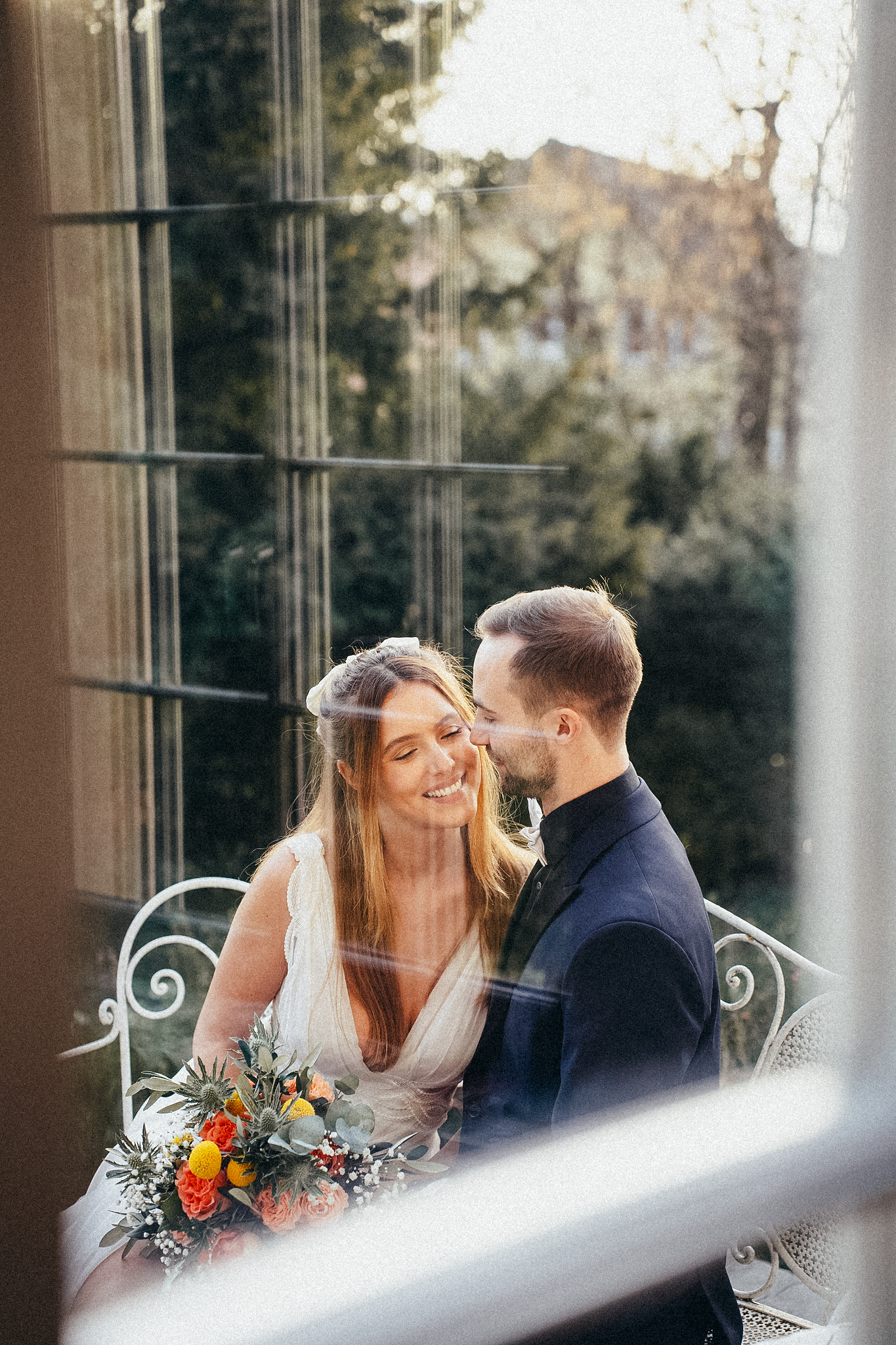 Groom kisses smiling bride on the cheek as they sit together, bride holding a colorful wedding bouquet.