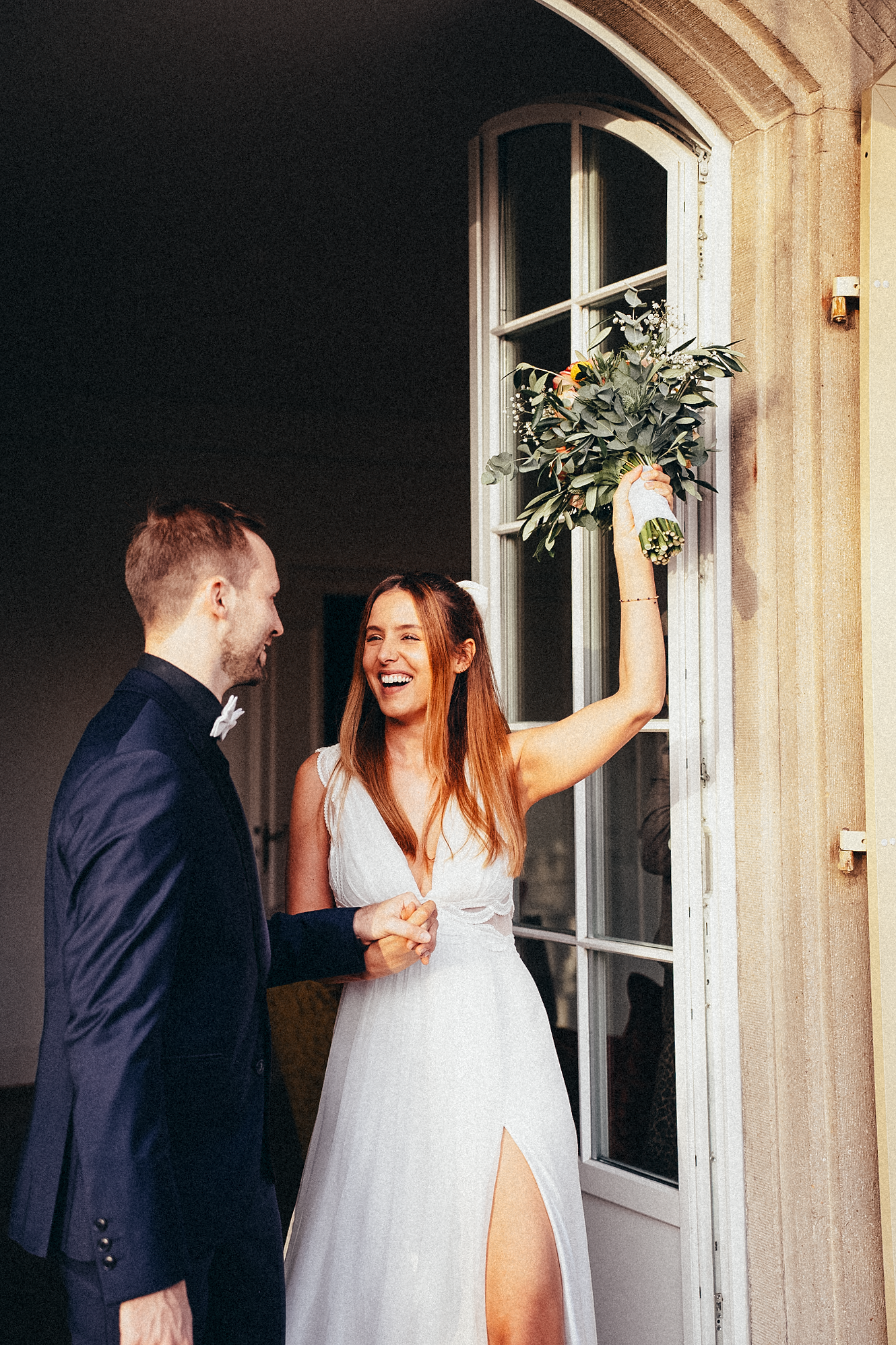 Laughing bride in white gown holds bouquet aloft while groom in navy suit holds her hand by elegant French doors.