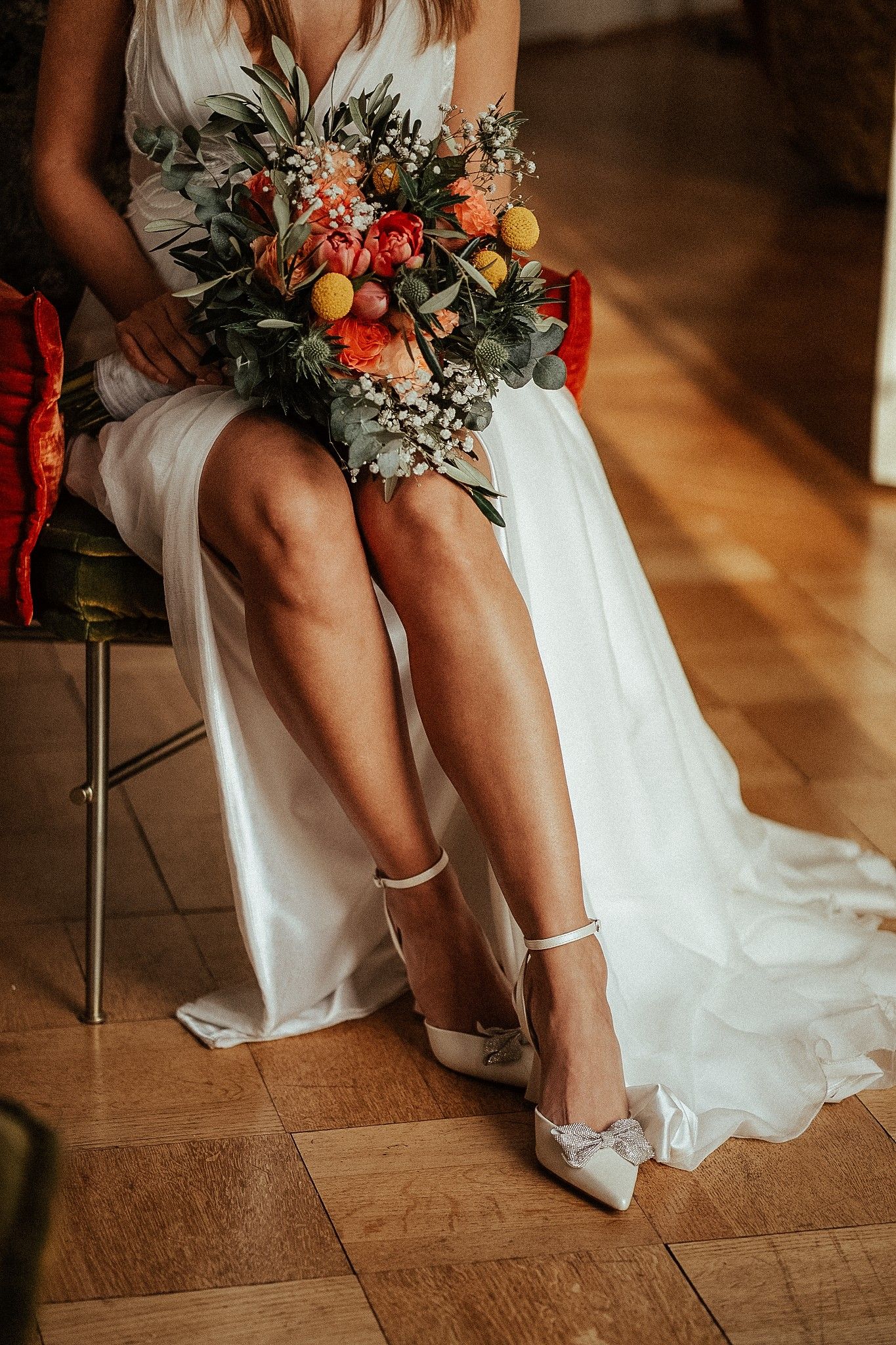 Bride in white gown holding colorful floral bouquet, seated with white bow heels on wooden floor.