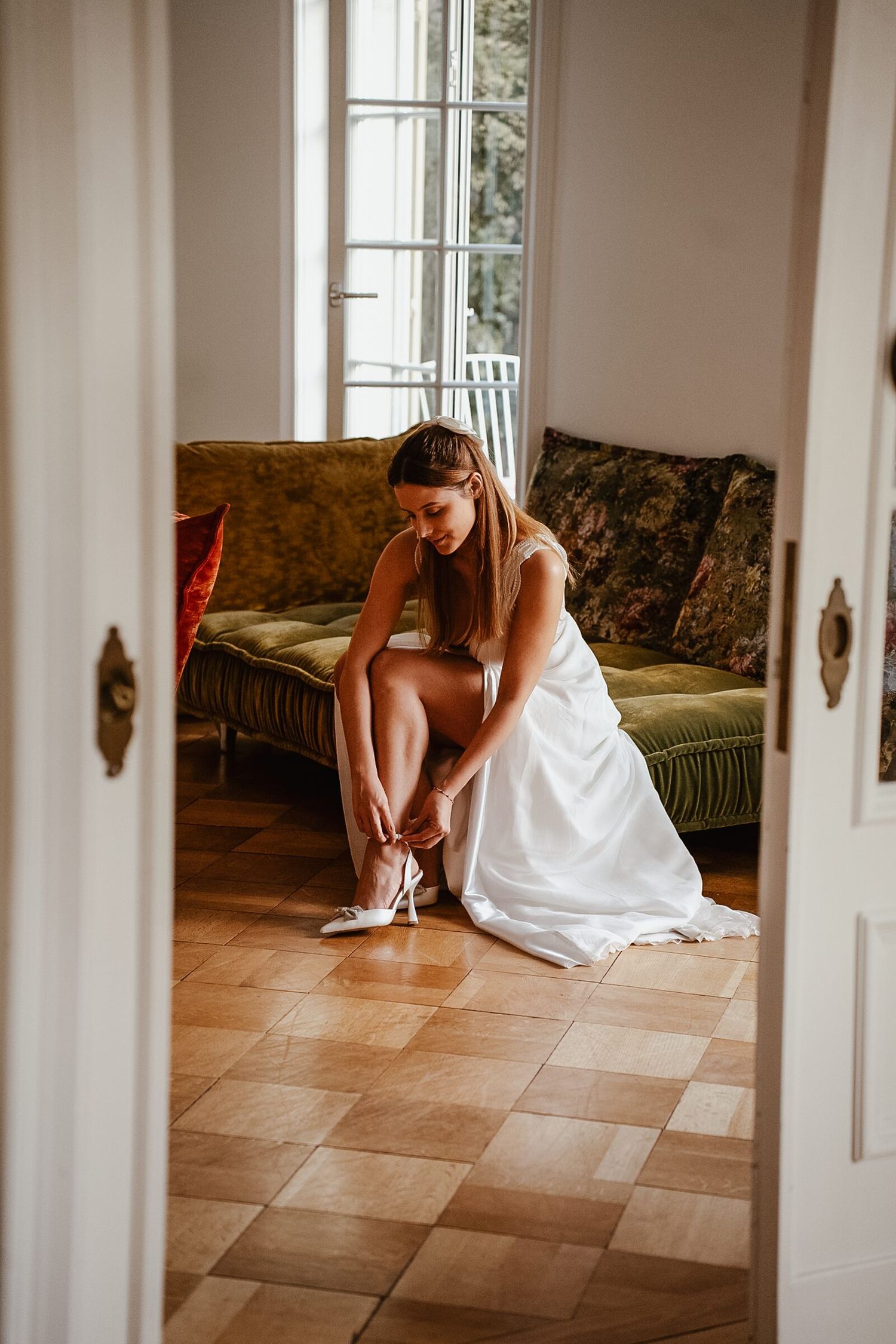 Bride in white wedding dress sitting on green sofa putting on heels in elegant room with large windows.