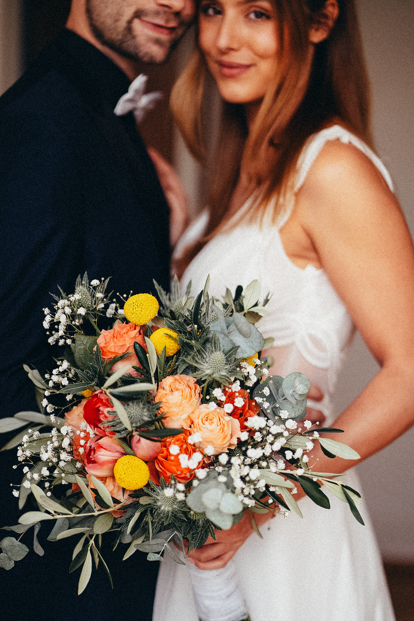 Bride in white dress holding colorful wedding bouquet with yellow, orange, and peach flowers with groom in background.
