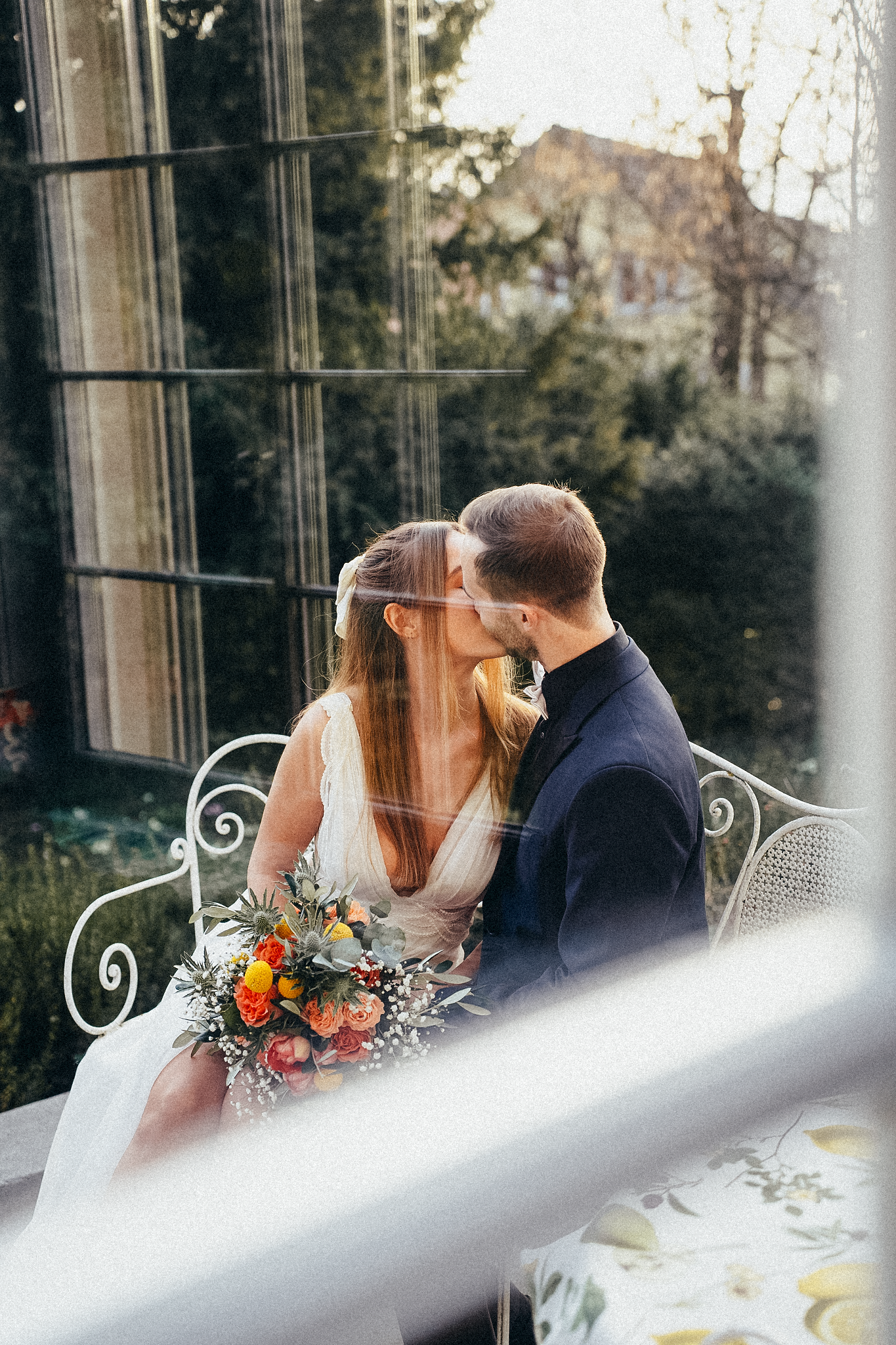 Bride and groom share a kiss on an elegant white bench outdoors, bride holding a vibrant floral bouquet.