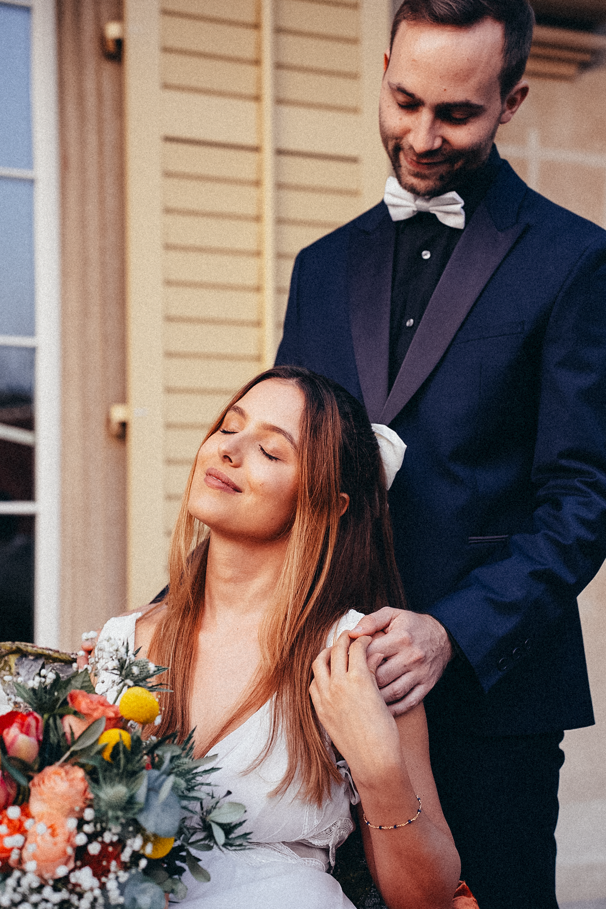 Groom in navy tuxedo with bow tie gently holds brides shoulders as she sits with eyes closed.
