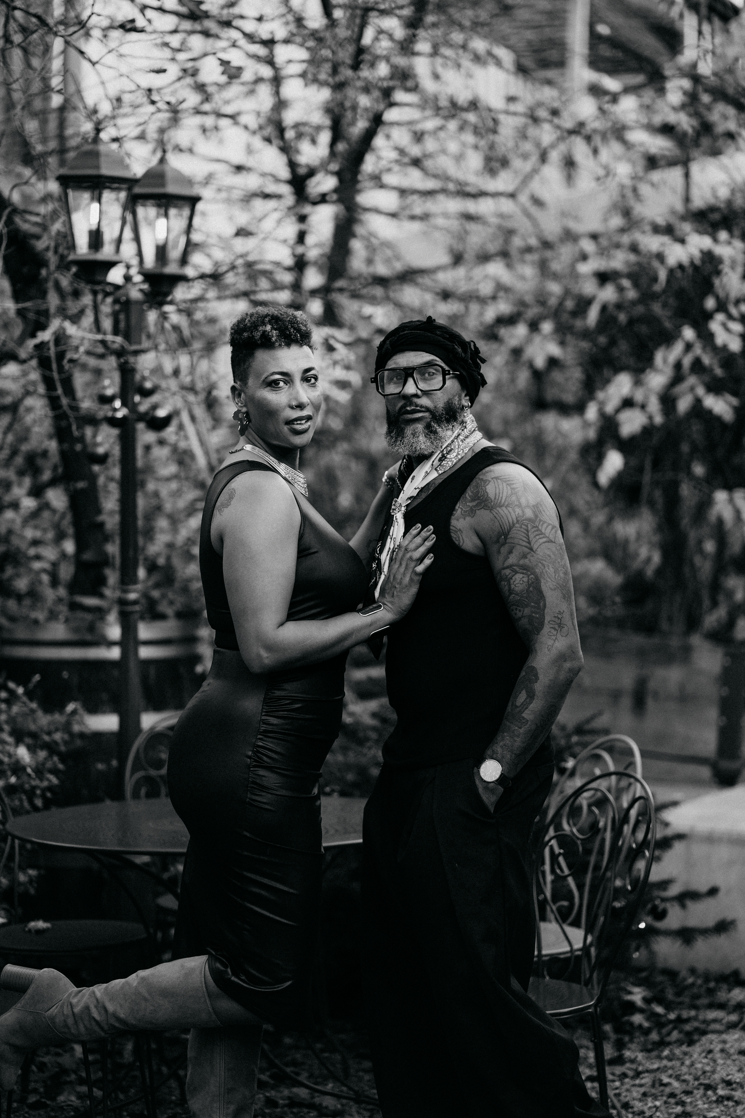 Black and white photo of a stylish couple posing outdoors in a garden setting with vintage lamp and lush foliage.