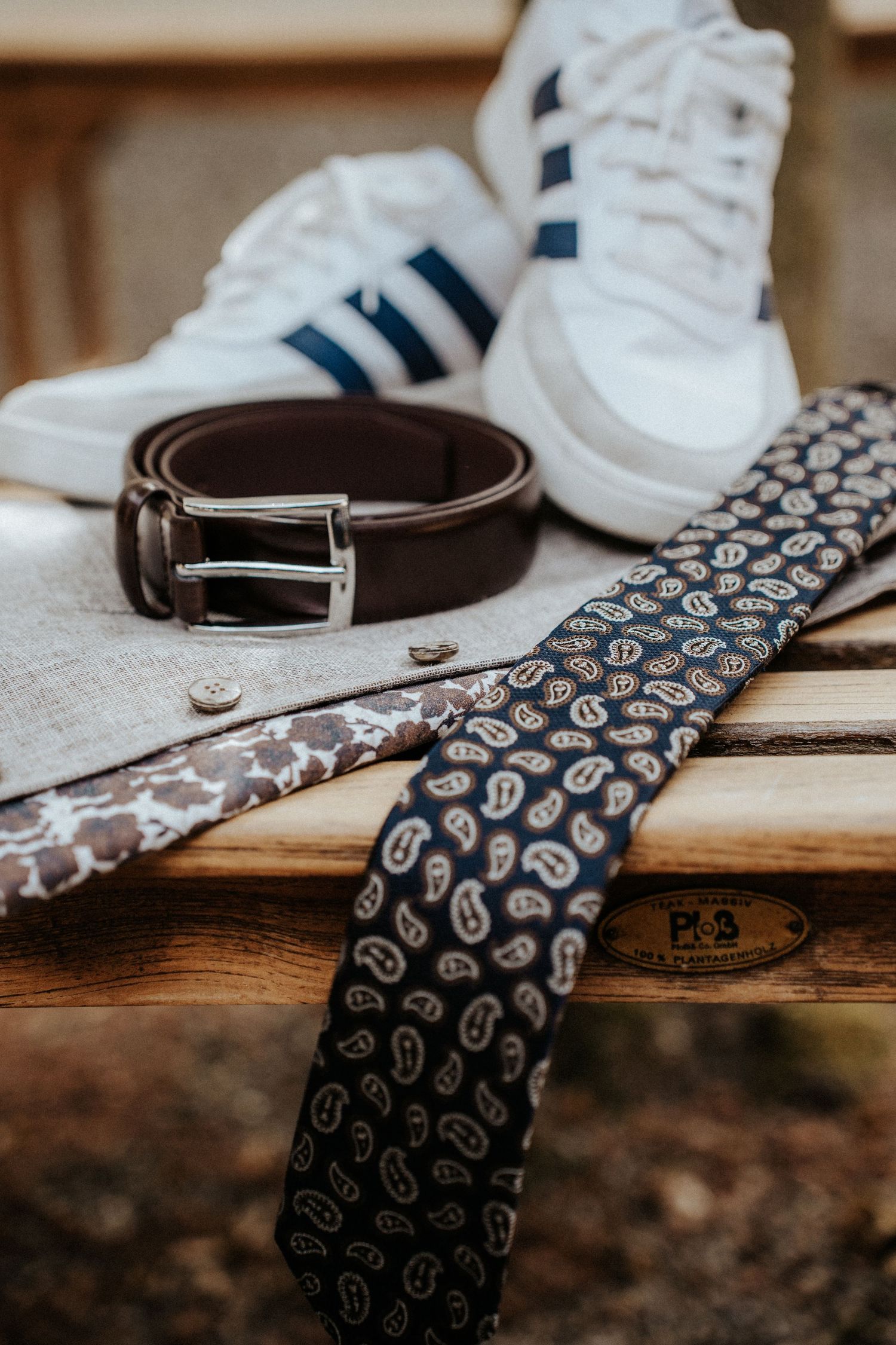 Men's accessories flat lay featuring a navy paisley tie, brown leather belt, pocket square, and white Adidas sneakers on wood.
