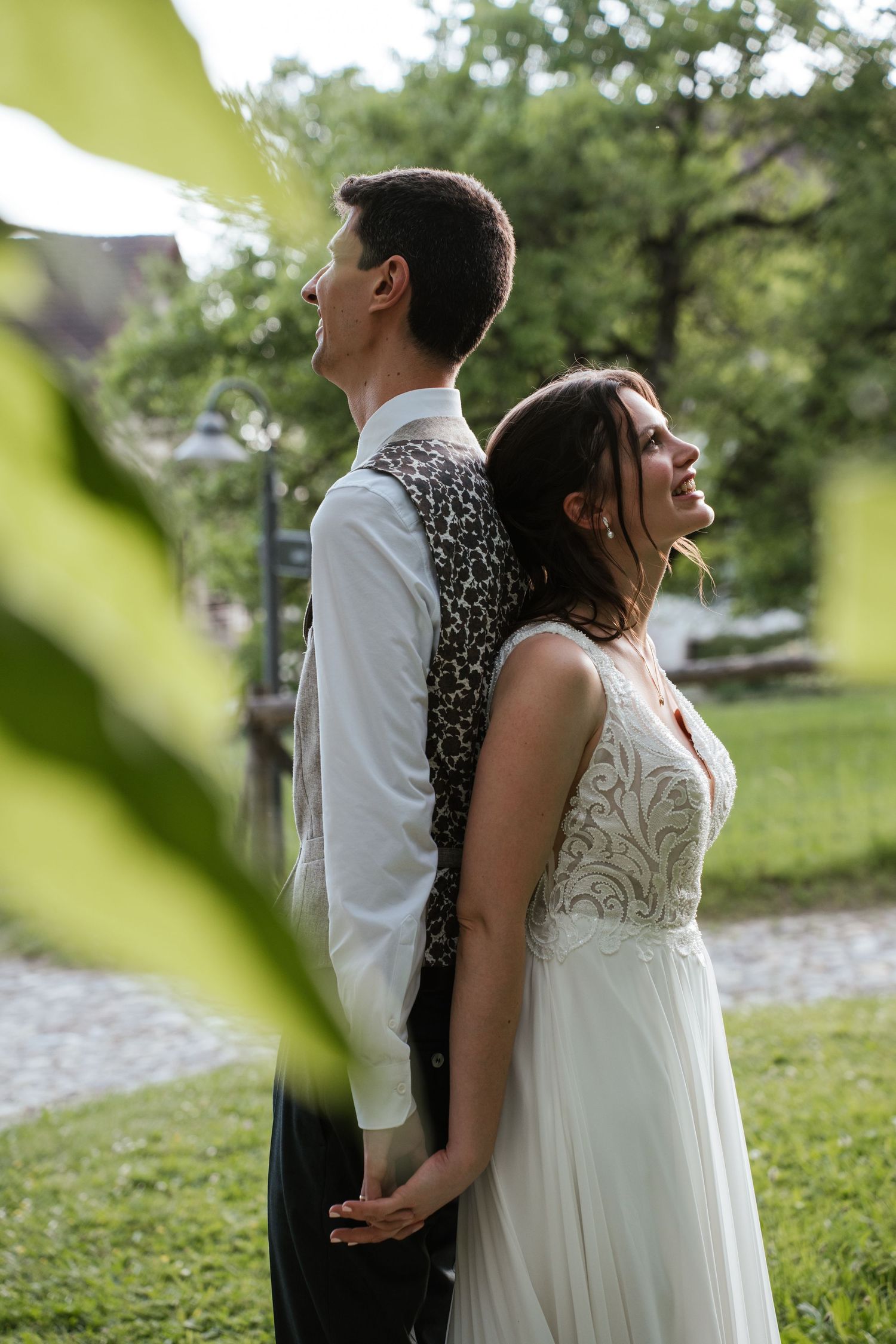 Bride and groom standing back-to-back in a lush garden, smiling joyfully in wedding attire outdoors.