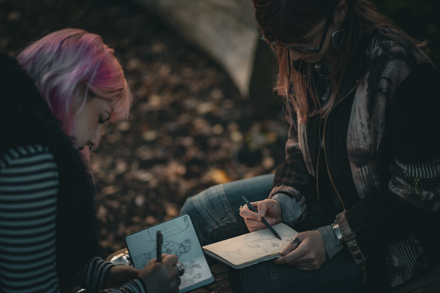 Two people sit outdoors, focused on sketching in notebooks. One has pink hair, the other wears a jacket. The setting is calm and creative.