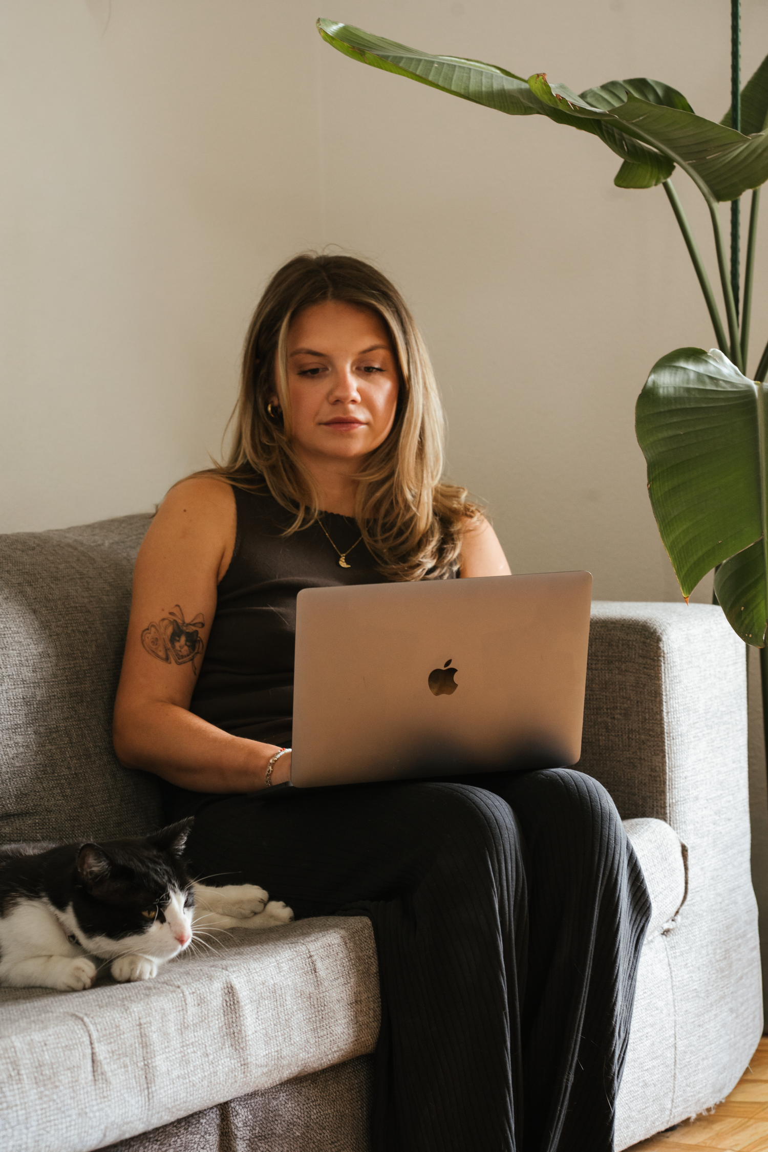 Woman with tattoos working on a MacBook laptop on a gray sofa with a black and white cat beside her.