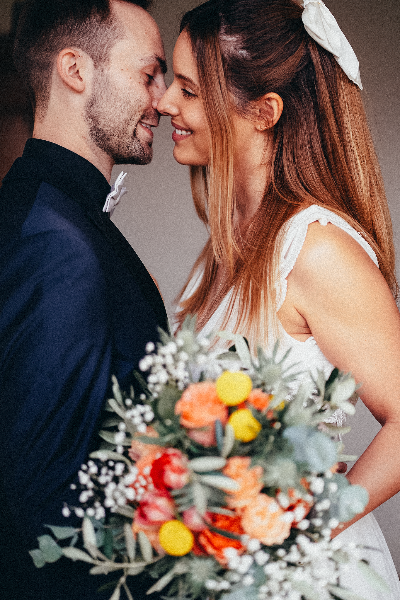 A smiling bride holding a colorful bouquet leans in close to her groom in a navy suit on their wedding day.