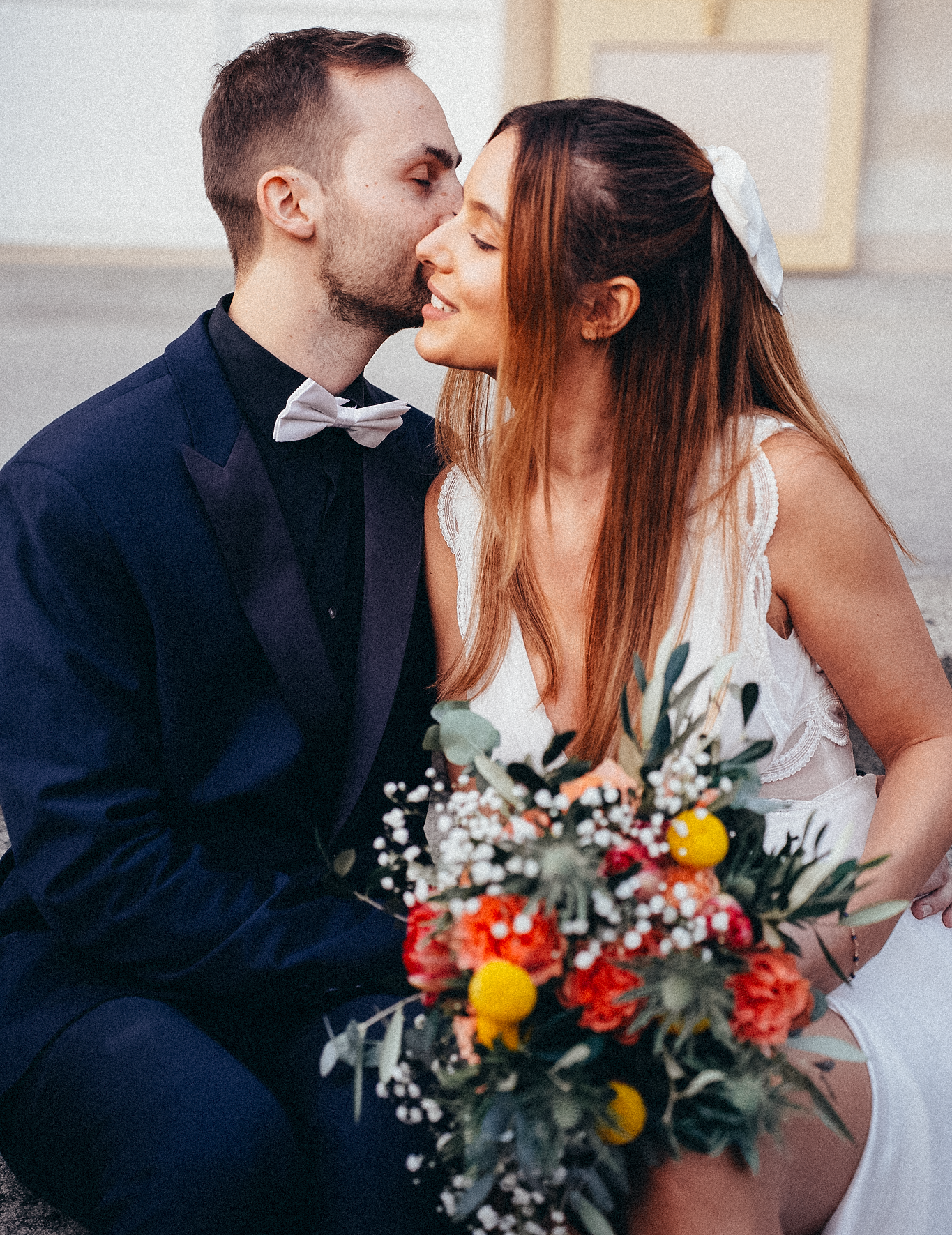 Smiling bride and groom sharing a tender kiss while seated outdoors, holding a vibrant wedding bouquet.