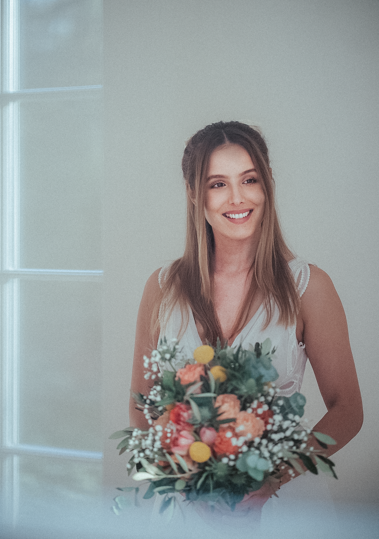 Smiling bride in white dress holding colorful spring bouquet with roses and greenery near bright window.
