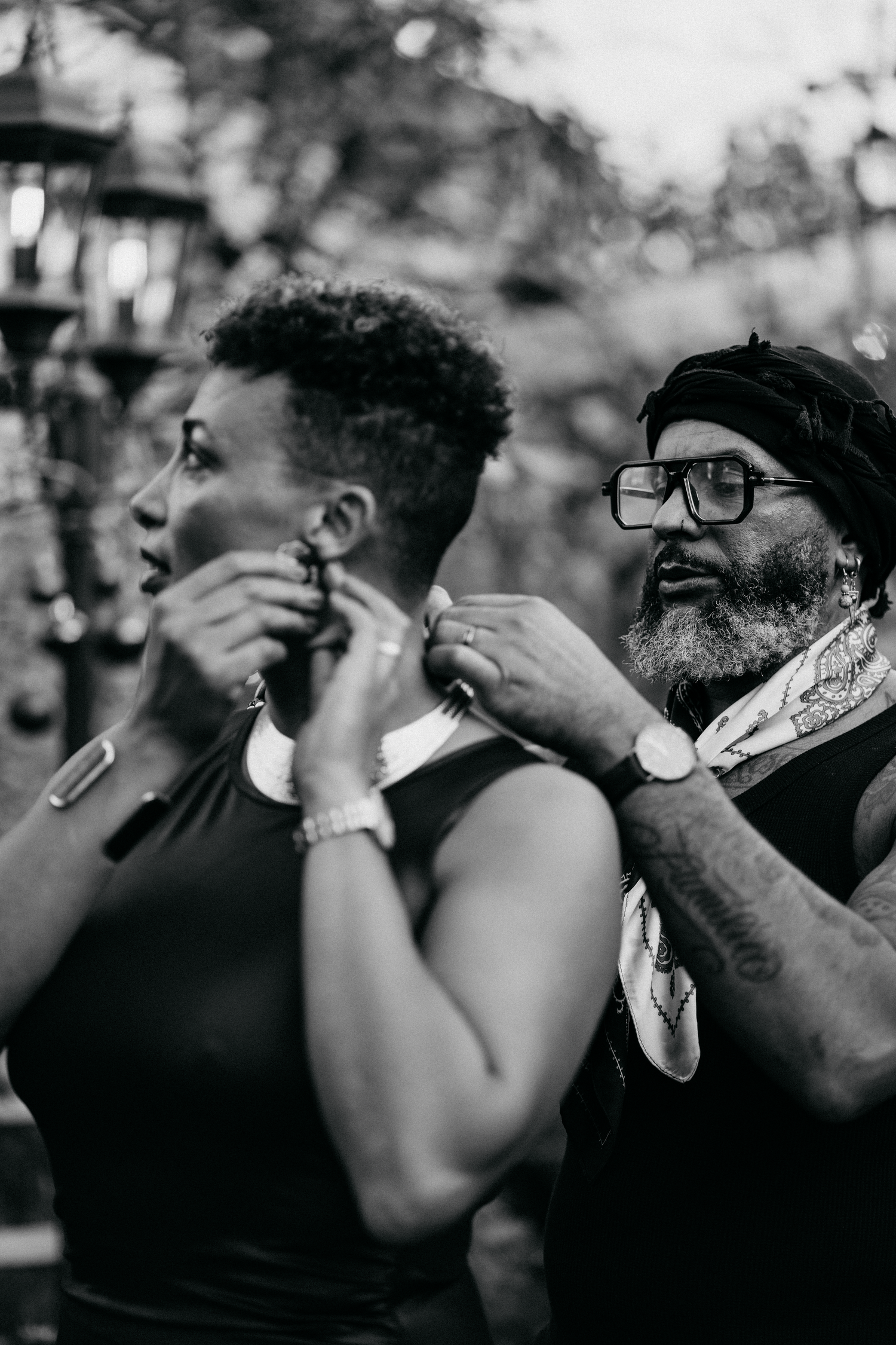 Black and white photo of a man helping a woman put on a necklace outdoors, both dressed elegantly.