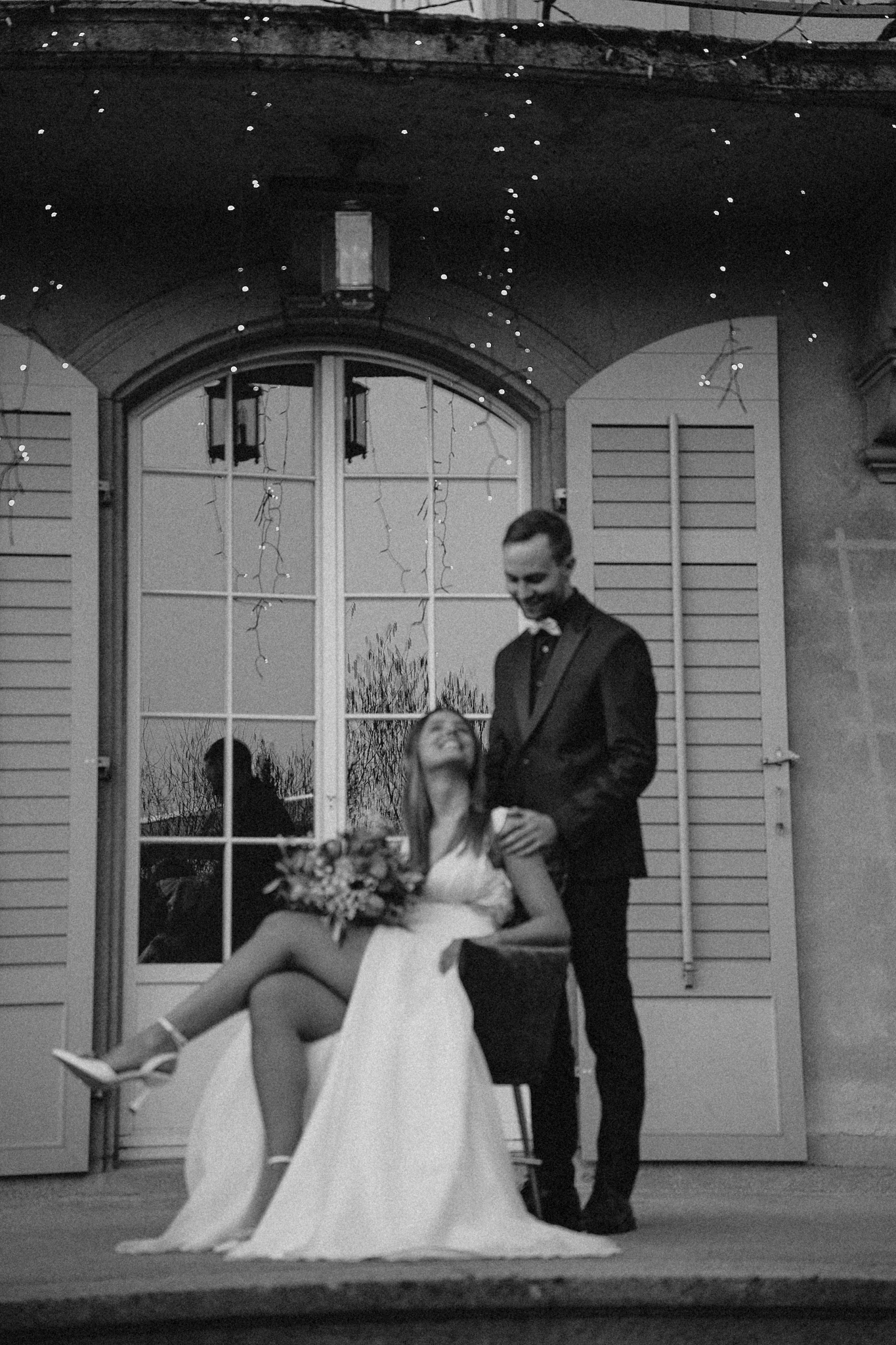 Black and white photo of bride sitting in chair leaning back while groom stands beside her outdoors.