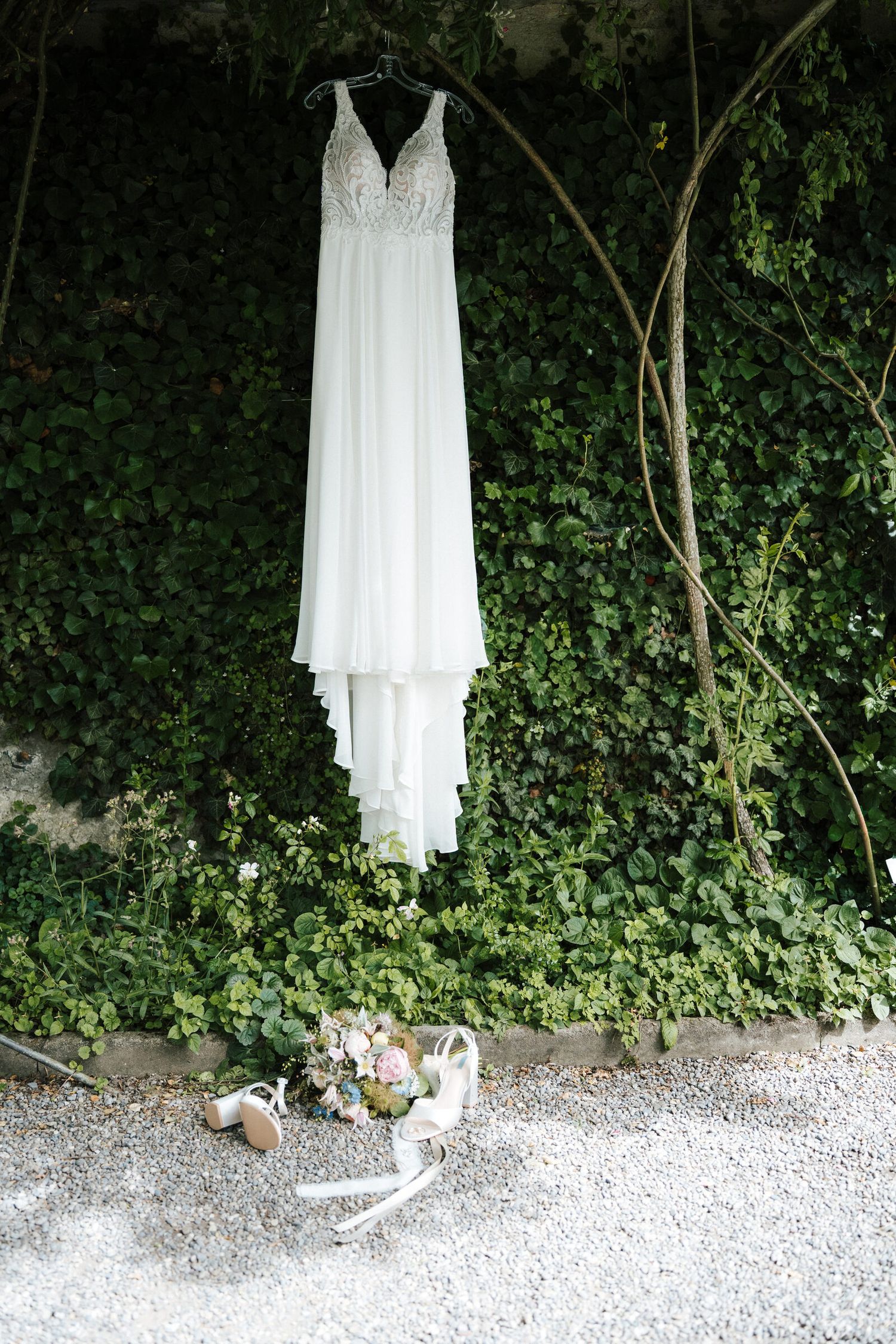 White lace wedding dress hanging on ivy wall with bridal heels and bouquet on gravel below.