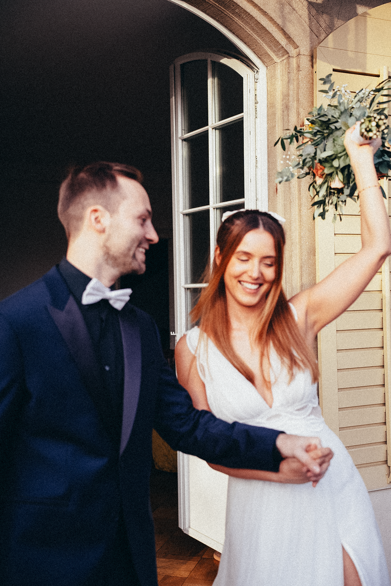 Joyful bride and groom holding hands outdoors, bride raising bouquet, groom in navy tuxedo, bride in white gown.