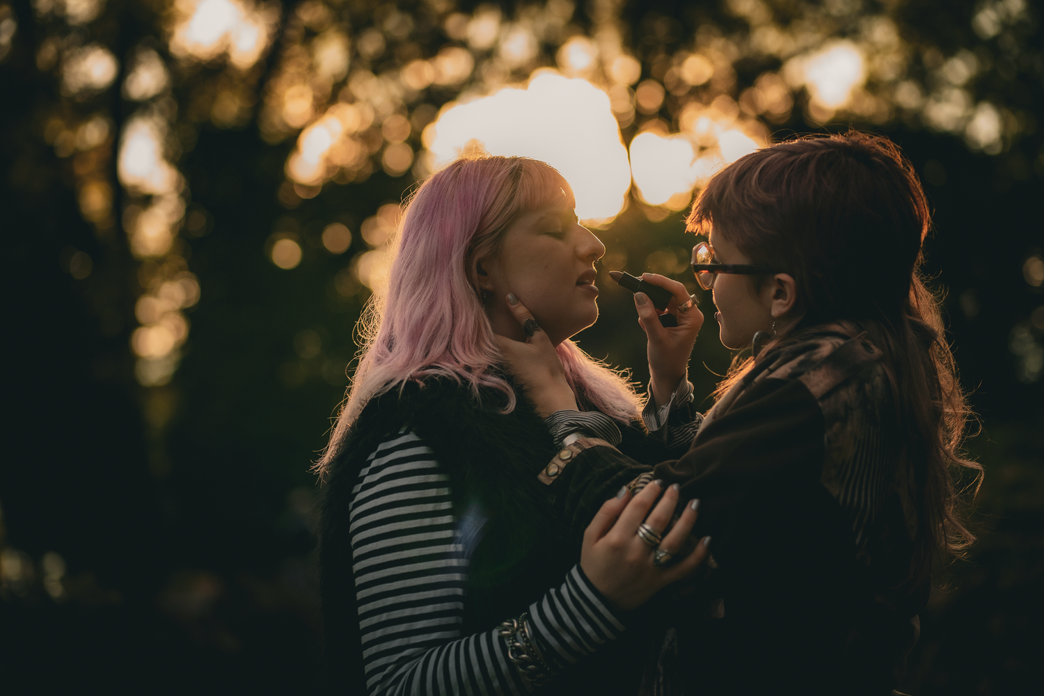 Two people are outdoors, one applying lipstick to the other. The warm, glowing sunset creates a bokeh effect