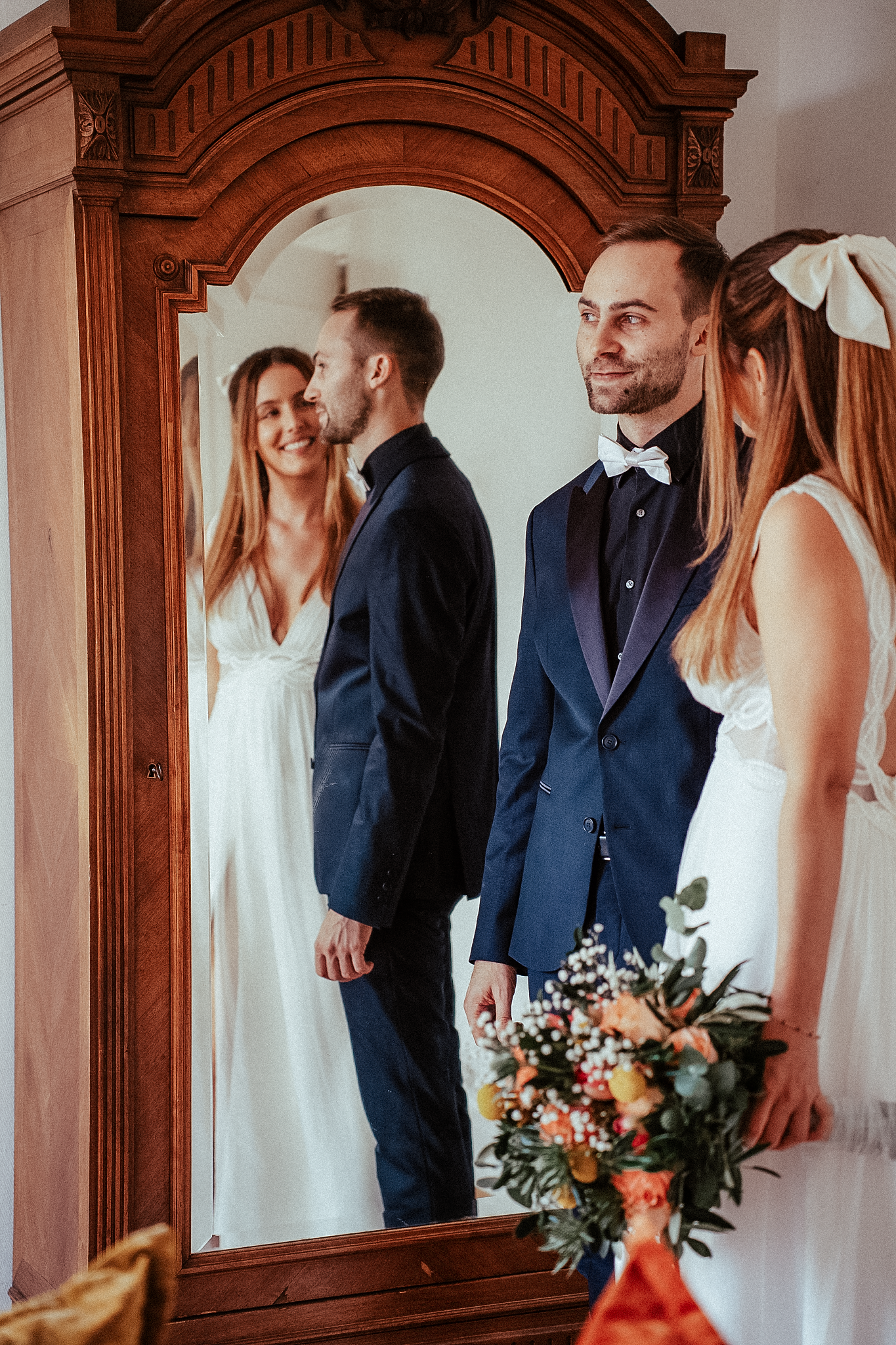 Wedding couple admires their reflection in ornate wooden mirror, bride in white gown holding bouquet.