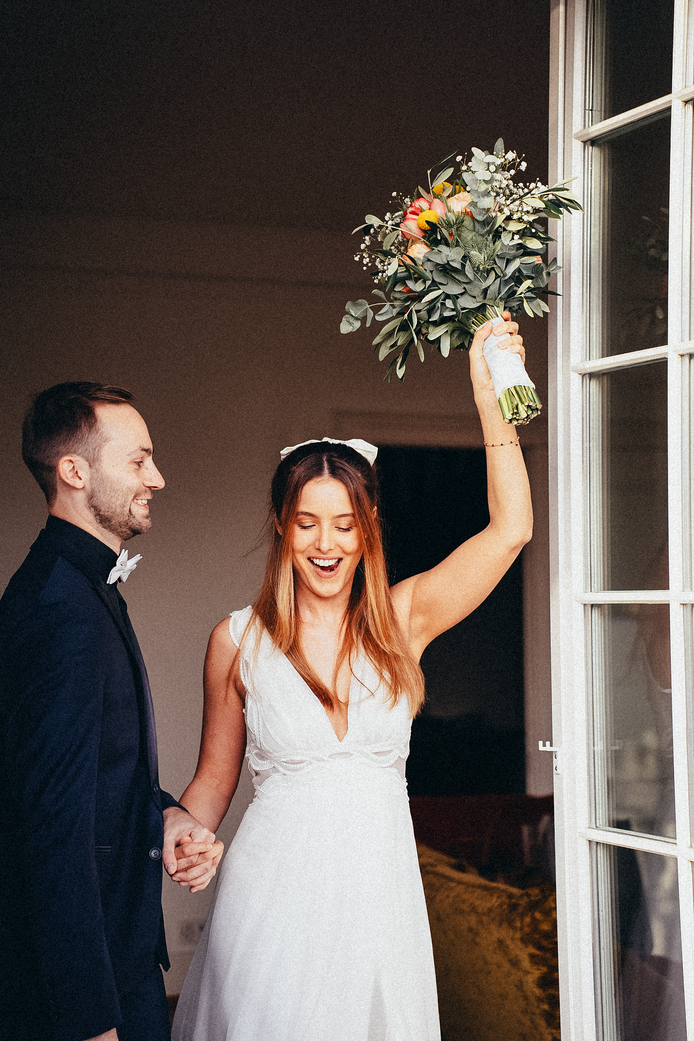 Joyful bride raises colorful bouquet while laughing groom holds her hand near elegant French doors.