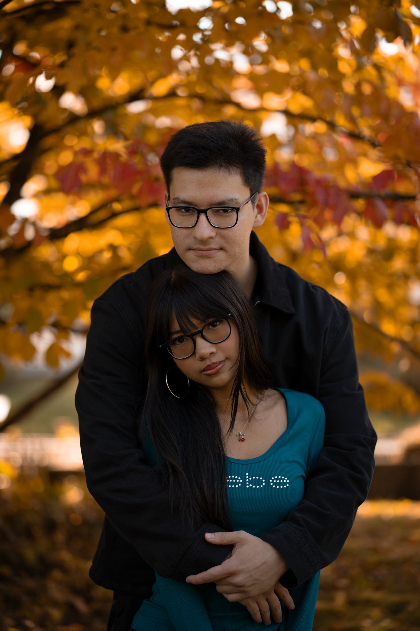Couple embracing outdoors in autumn park with vibrant orange and yellow foliage, both wearing glasses and dark jackets.