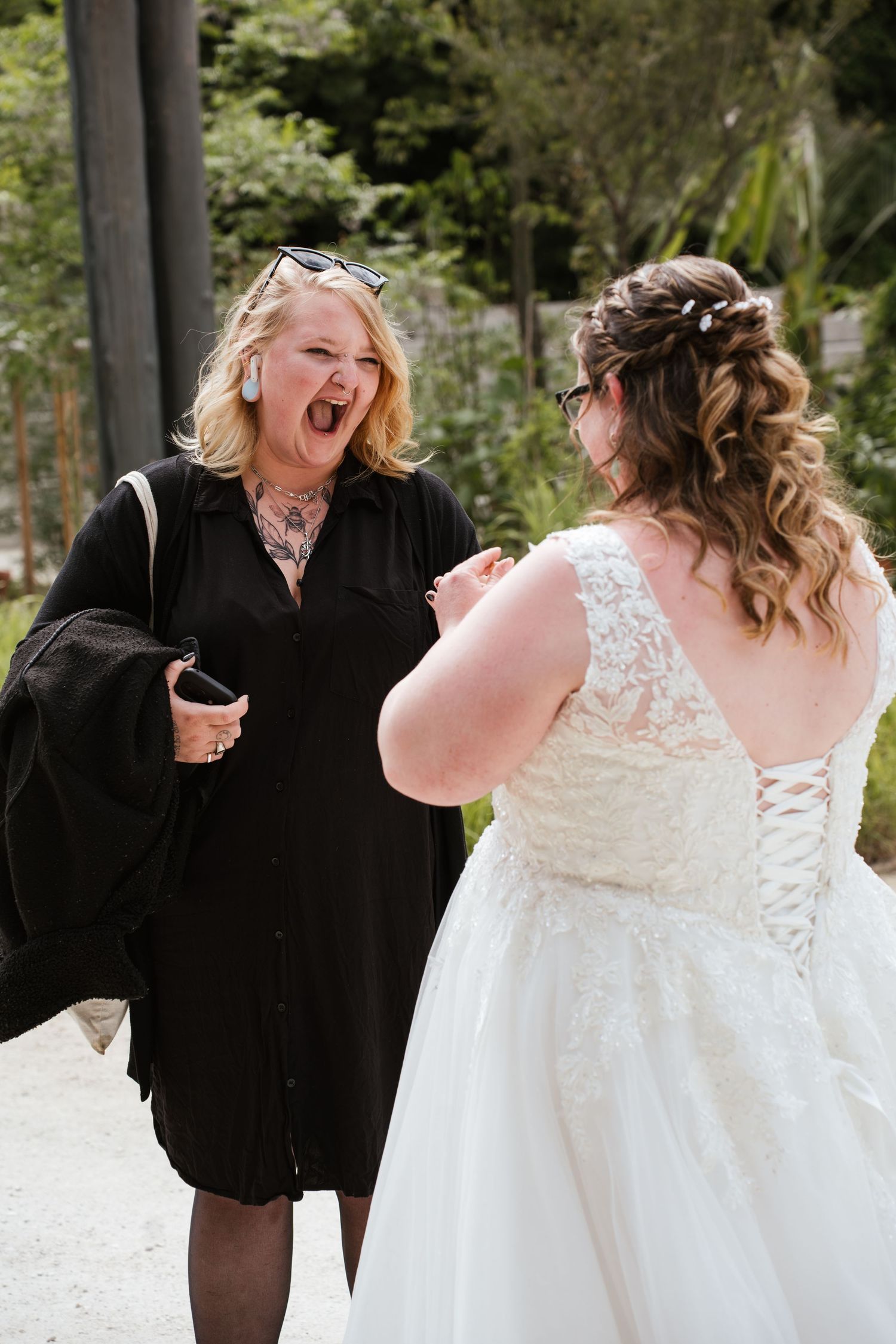 A bride in a lace gown shares a joyful moment with a woman in black at an outdoor wedding venue.