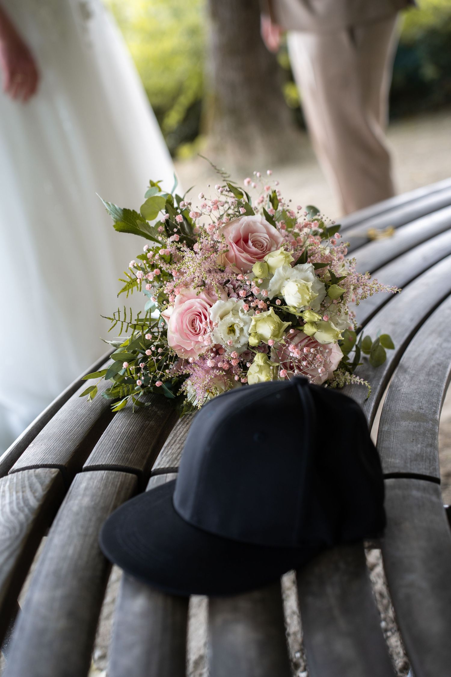 Wedding bouquet of pink roses and white flowers resting on a wooden bench beside a black cap.