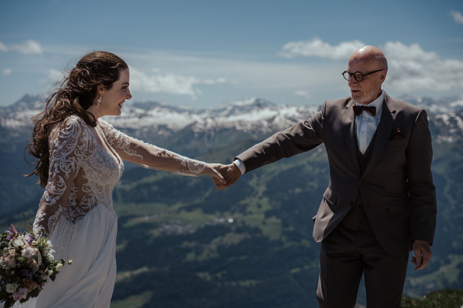 Bride and groom holding hands on a mountain summit with stunning snow-capped Alps in the background.
