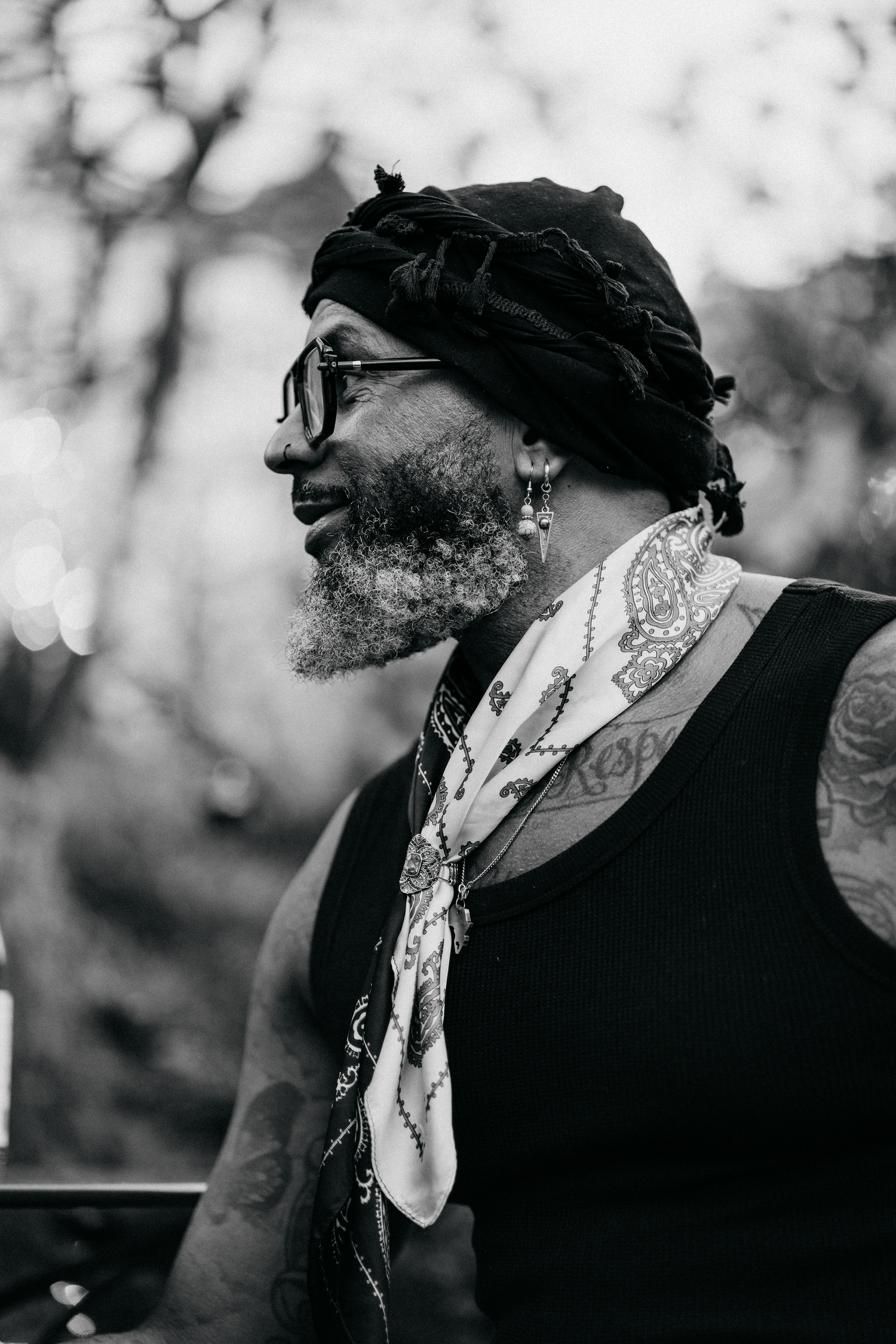 Black and white portrait of tattooed man with beard, dreadlocks, sunglasses, and patterned scarf outdoors.