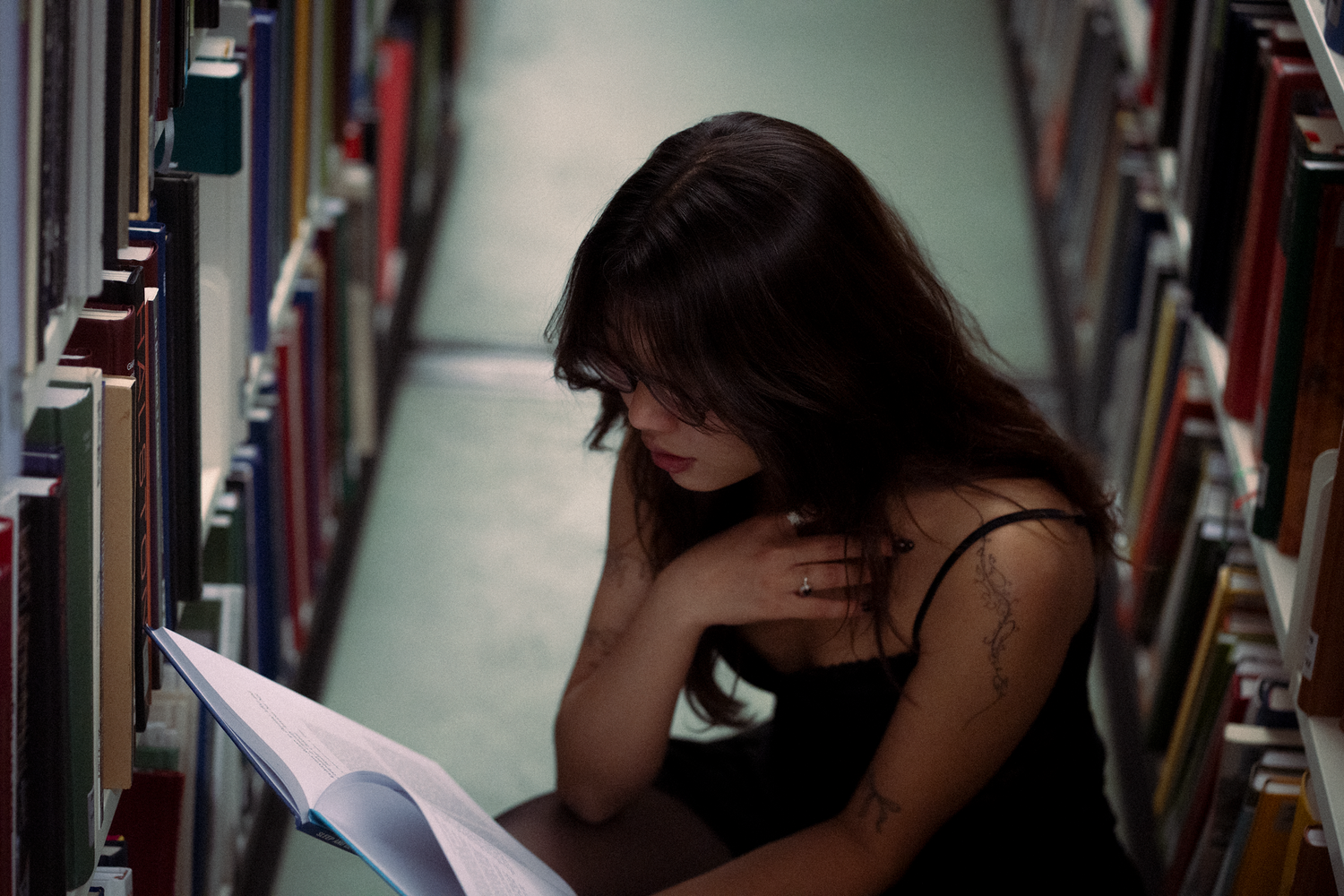 Young woman with tattoo reading a book while sitting on the floor between library bookshelves.