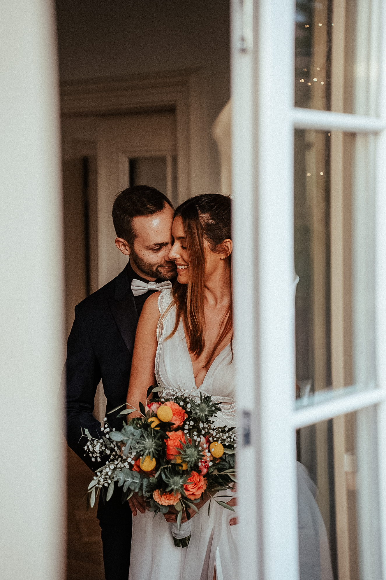 Bride and groom share an intimate embrace framed by elegant white French doors, holding a vibrant bouquet.