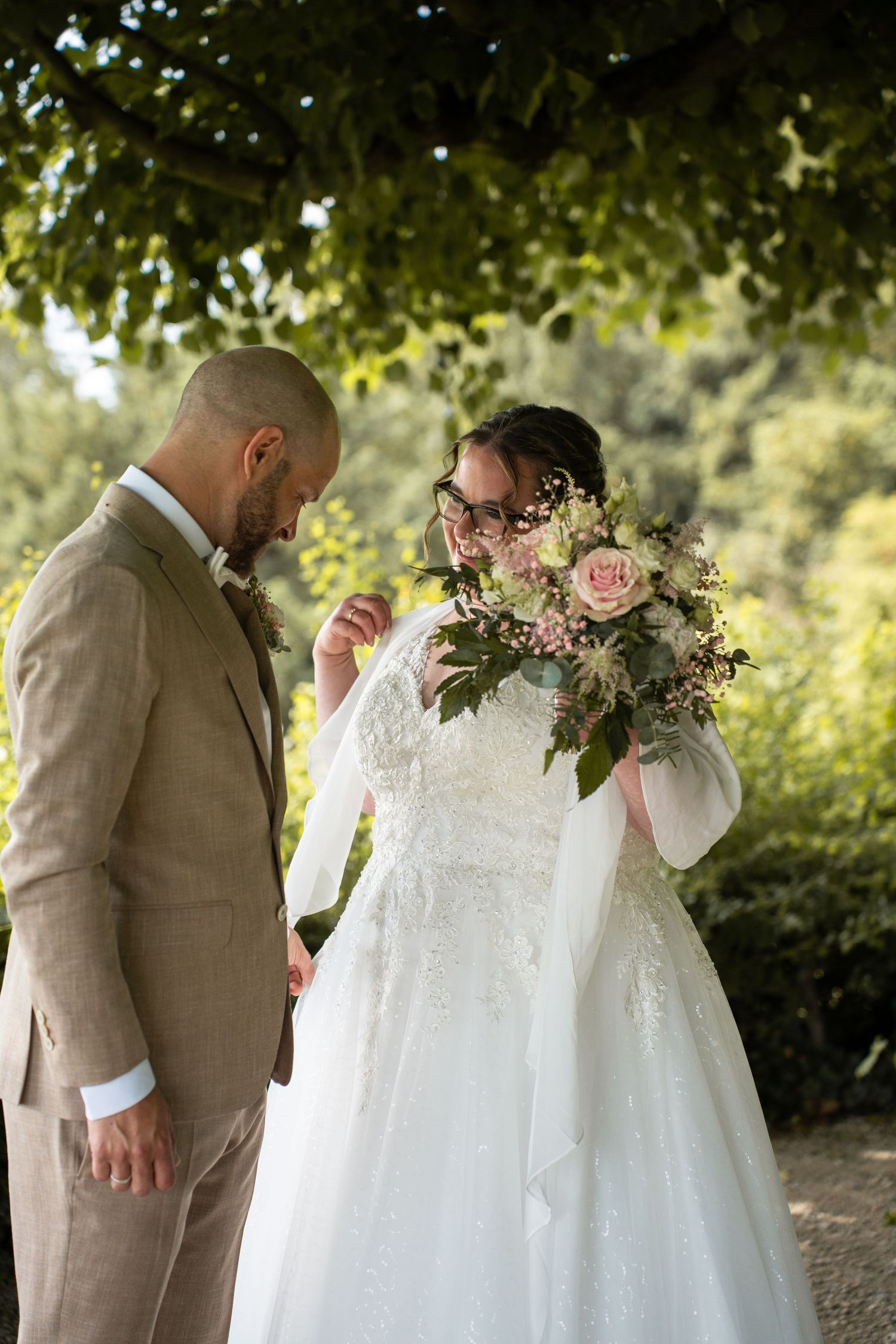 A bride in a white lace gown holds a pink floral bouquet beside a groom in a tan suit outdoors.