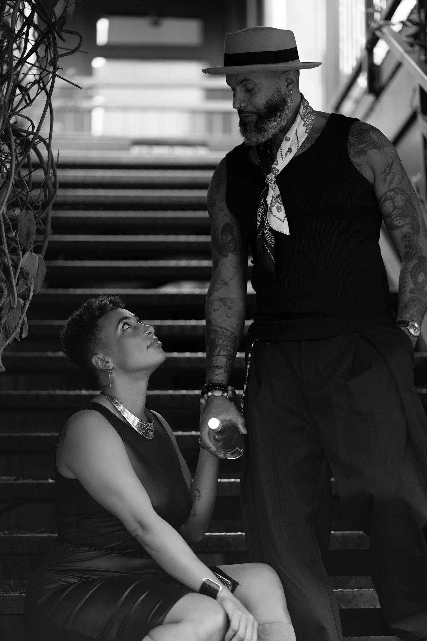 Black and white photo of tattooed man in hat standing on stairs looking down at woman seated below him.