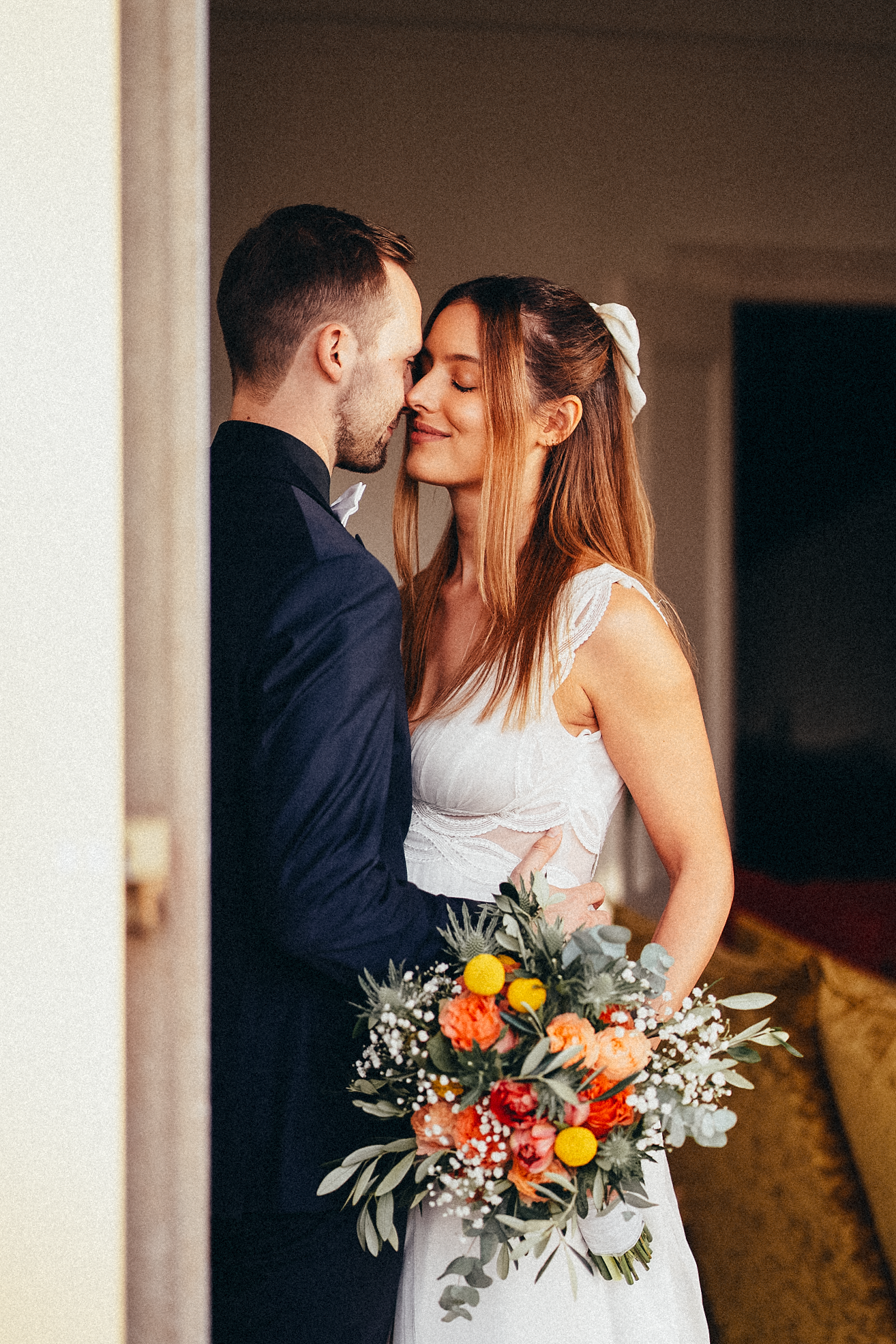 A bride and groom share a tender moment, she holds a colorful bouquet of orange and yellow flowers in a white dress.