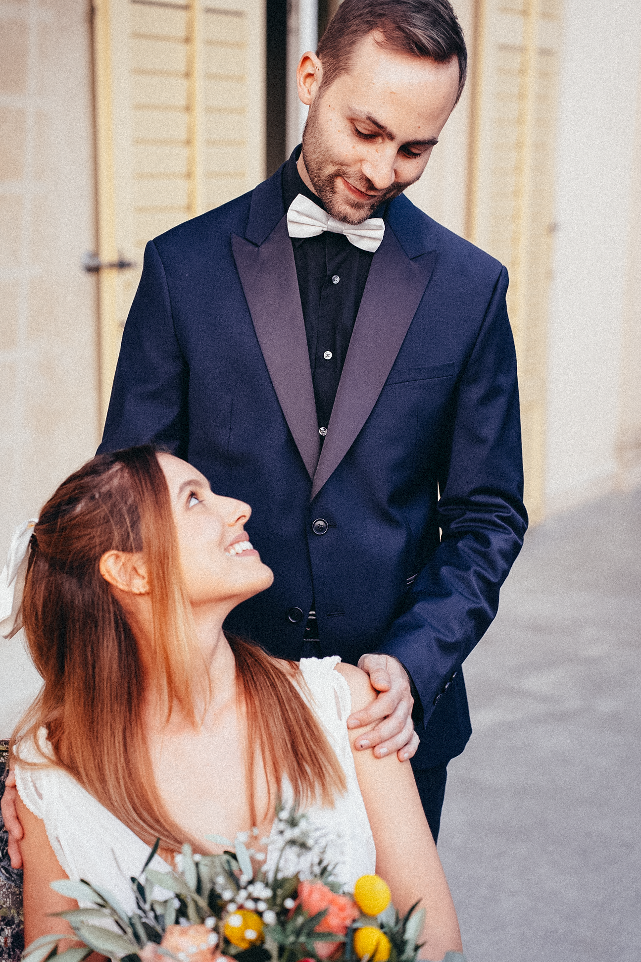 Bride looks up lovingly at groom in navy tuxedo while holding colorful bouquet outdoors.