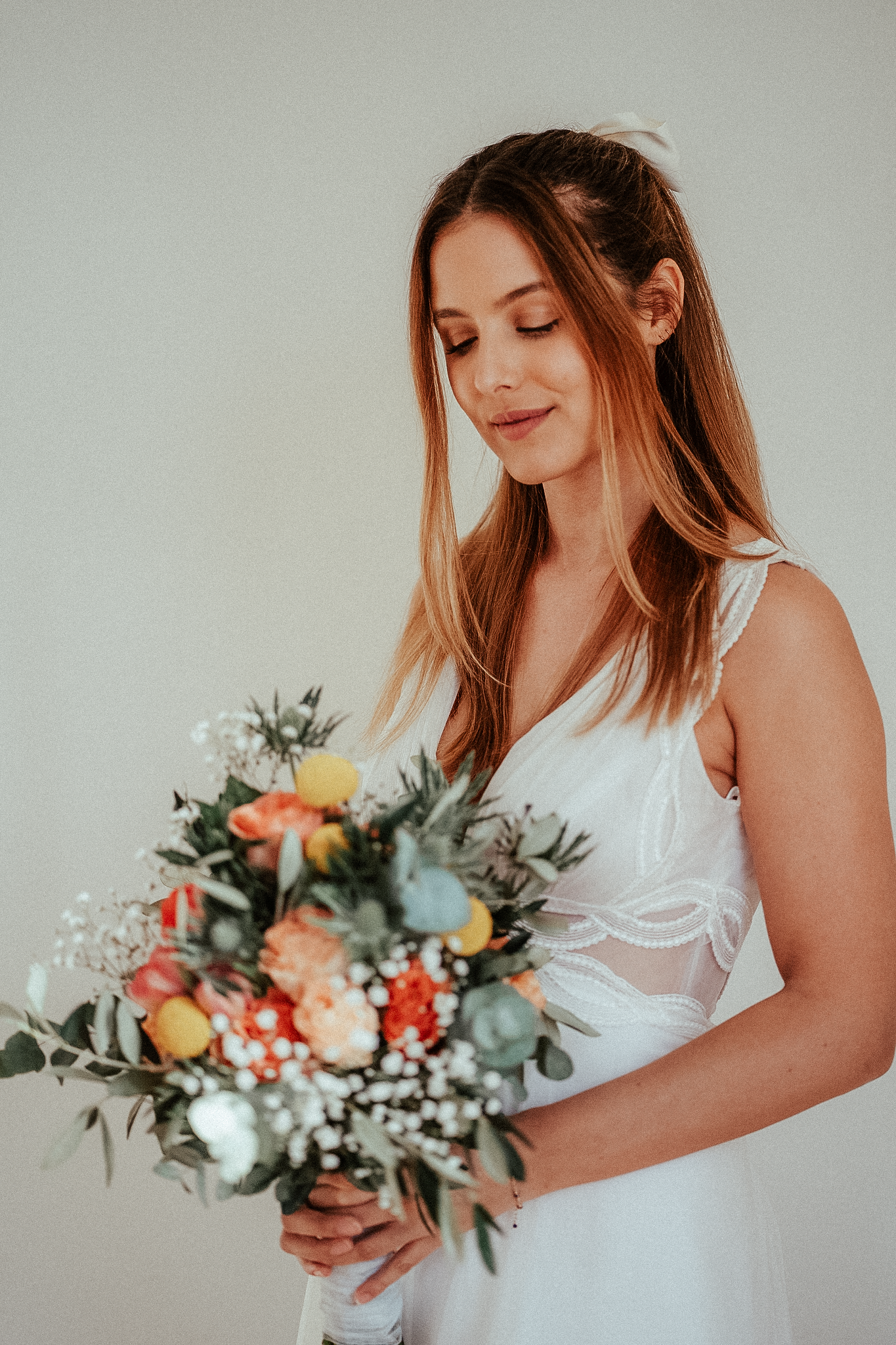 Bride in white gown holding colorful bouquet with roses, baby's breath, and eucalyptus, smiling softly.