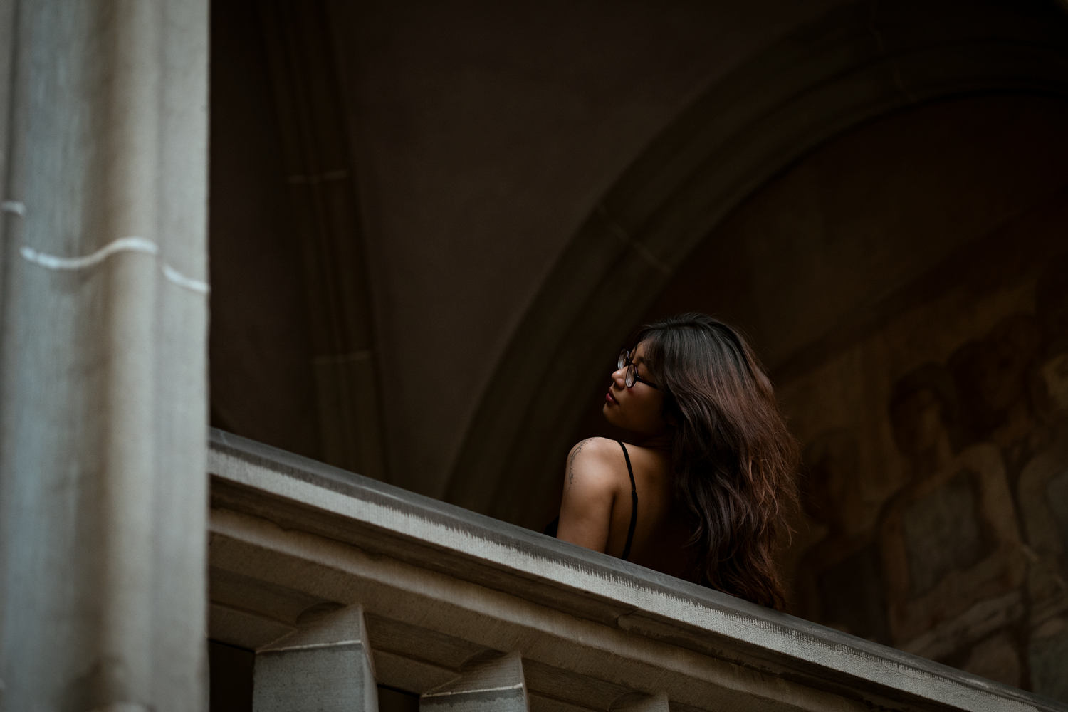 A young woman with long dark hair stands on a grand stone balcony with classical columns in a historic building.