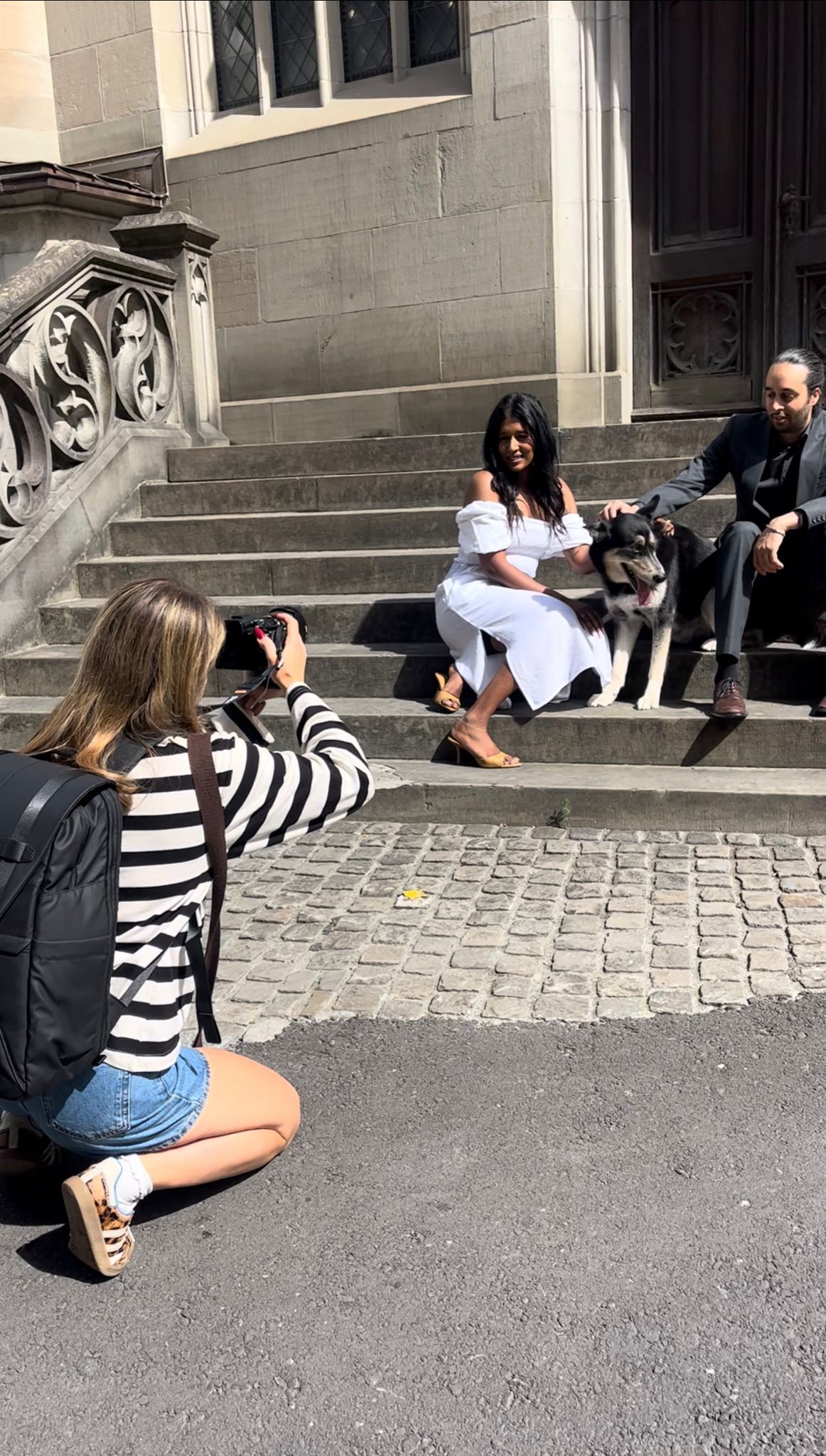 Woman photographer kneeling on cobblestone street photographing couple with dog on steps.