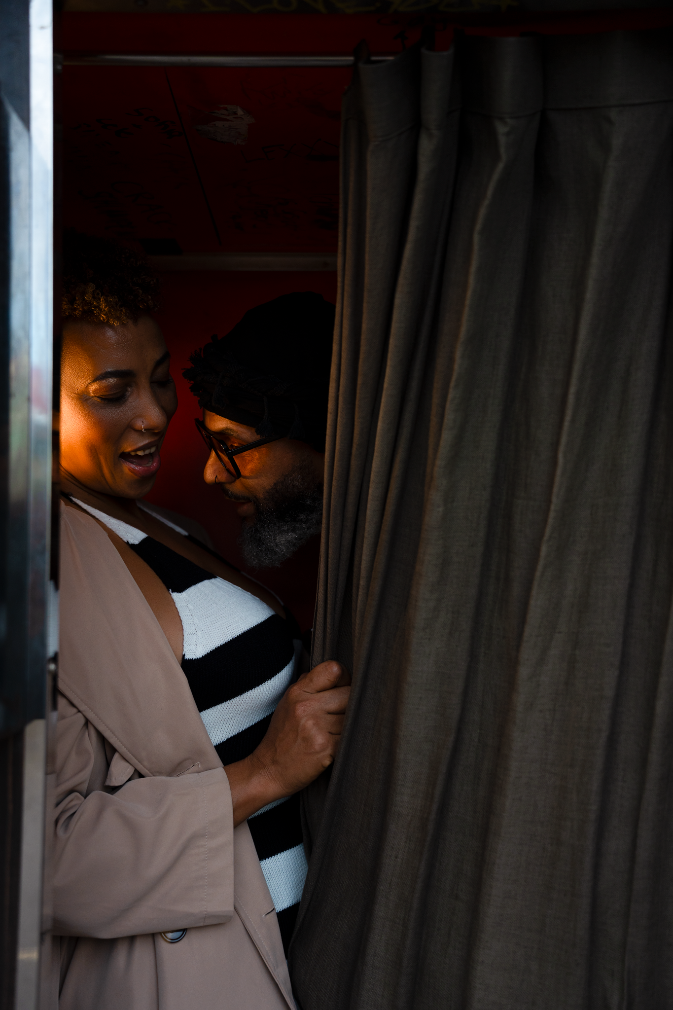 Couple sharing an intimate moment peeking through a gray curtain in a warmly lit red booth.